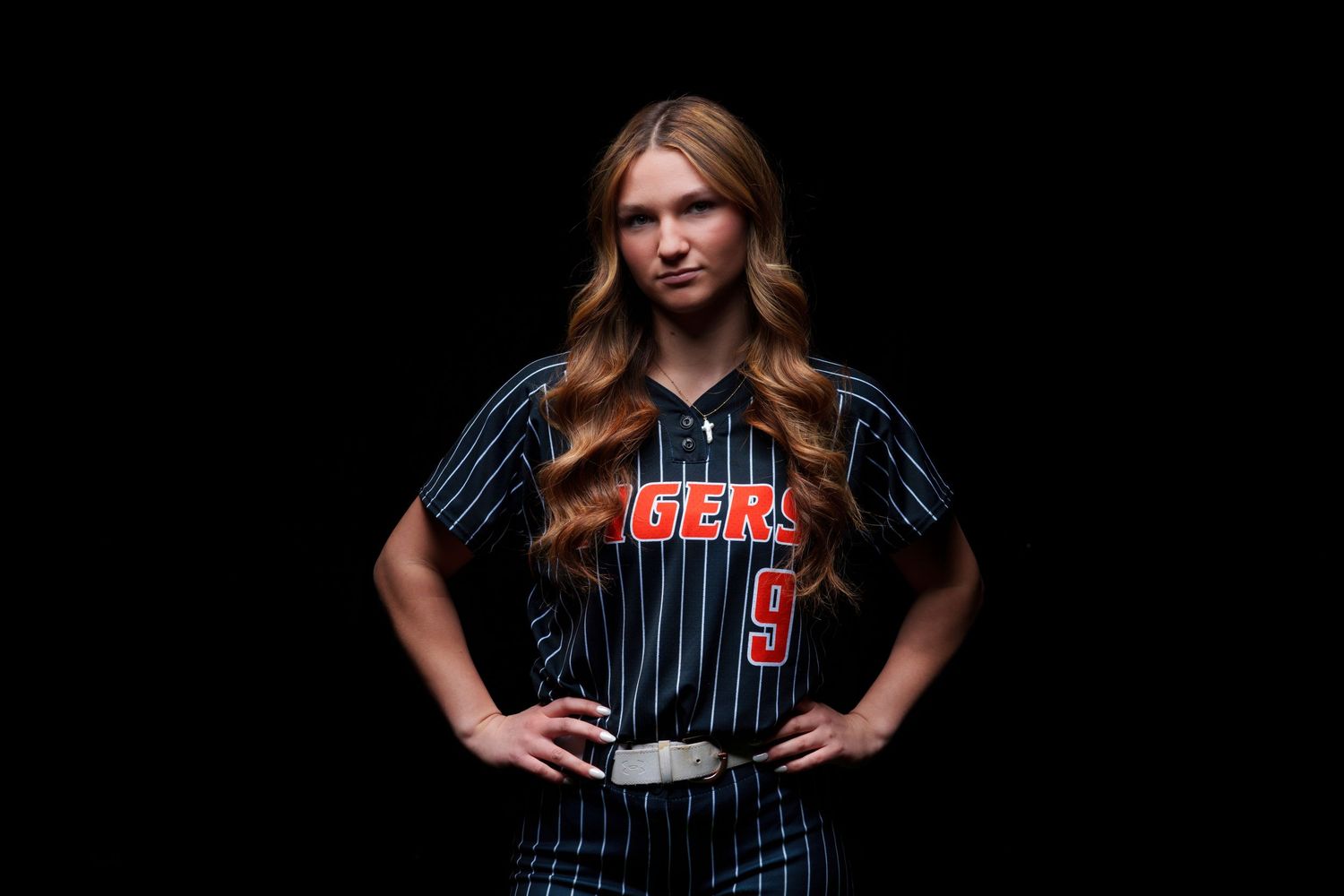 Young female softball player in Tigers pinstripe uniform posing confidently against dark background.