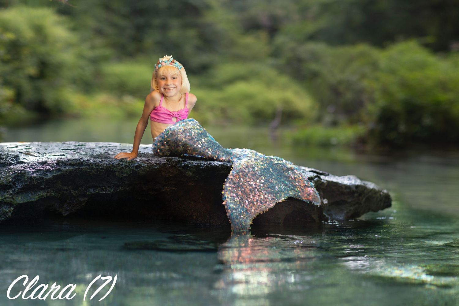 A mermaid tail sparkles in the sunlight as someone poses on a rock by a tranquil lake surrounded by trees.