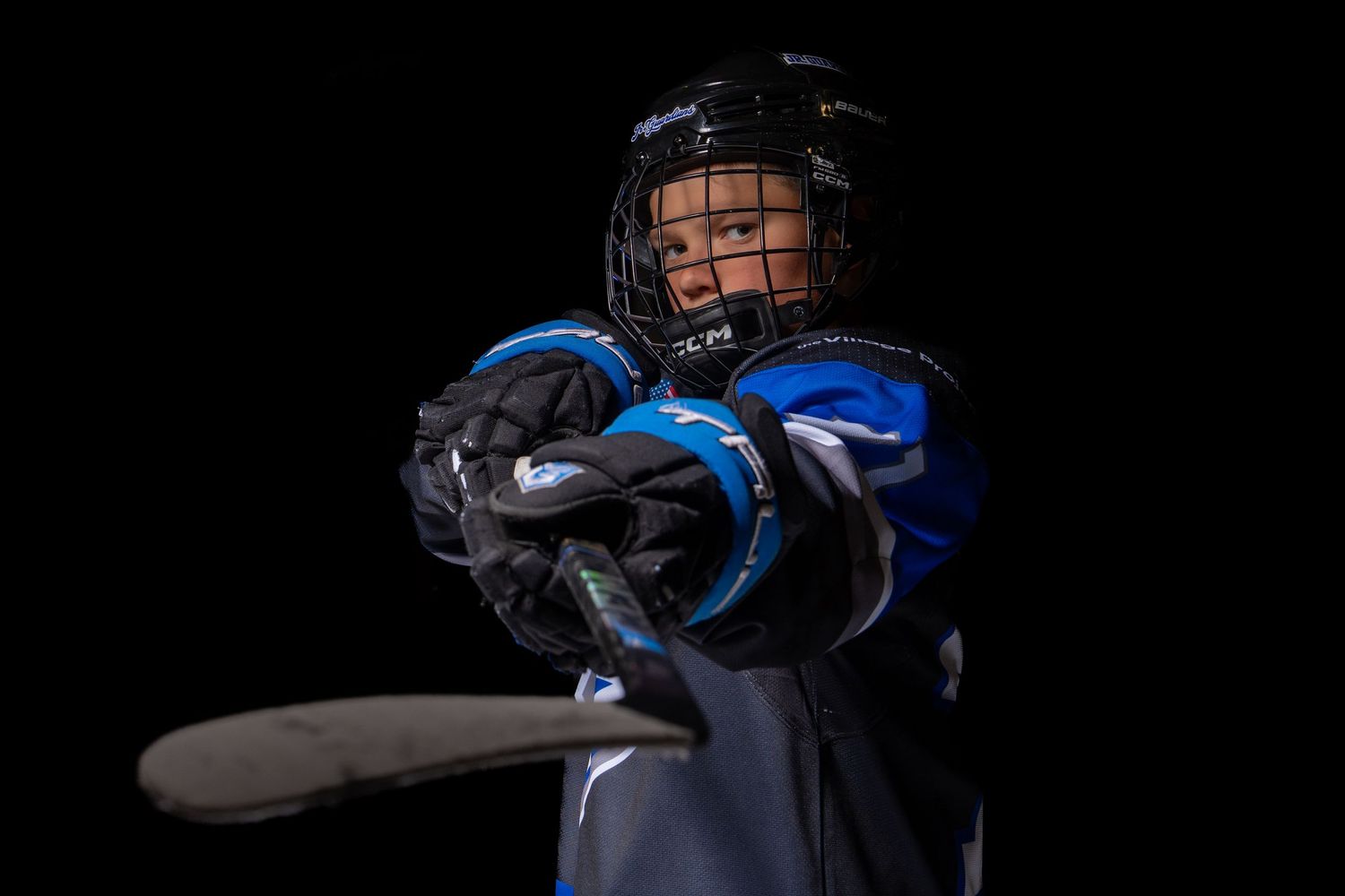 Youth hockey player in blue jersey and protective gear poses with stick against dark background.