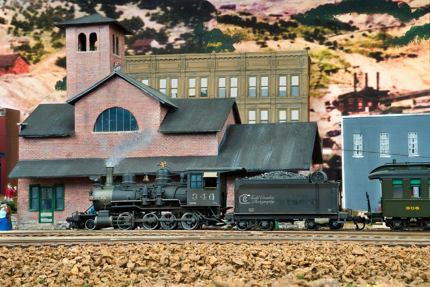 A vintage train yard scene with multiple locomotives and railcars against a backdrop of industrial buildings at sunset.