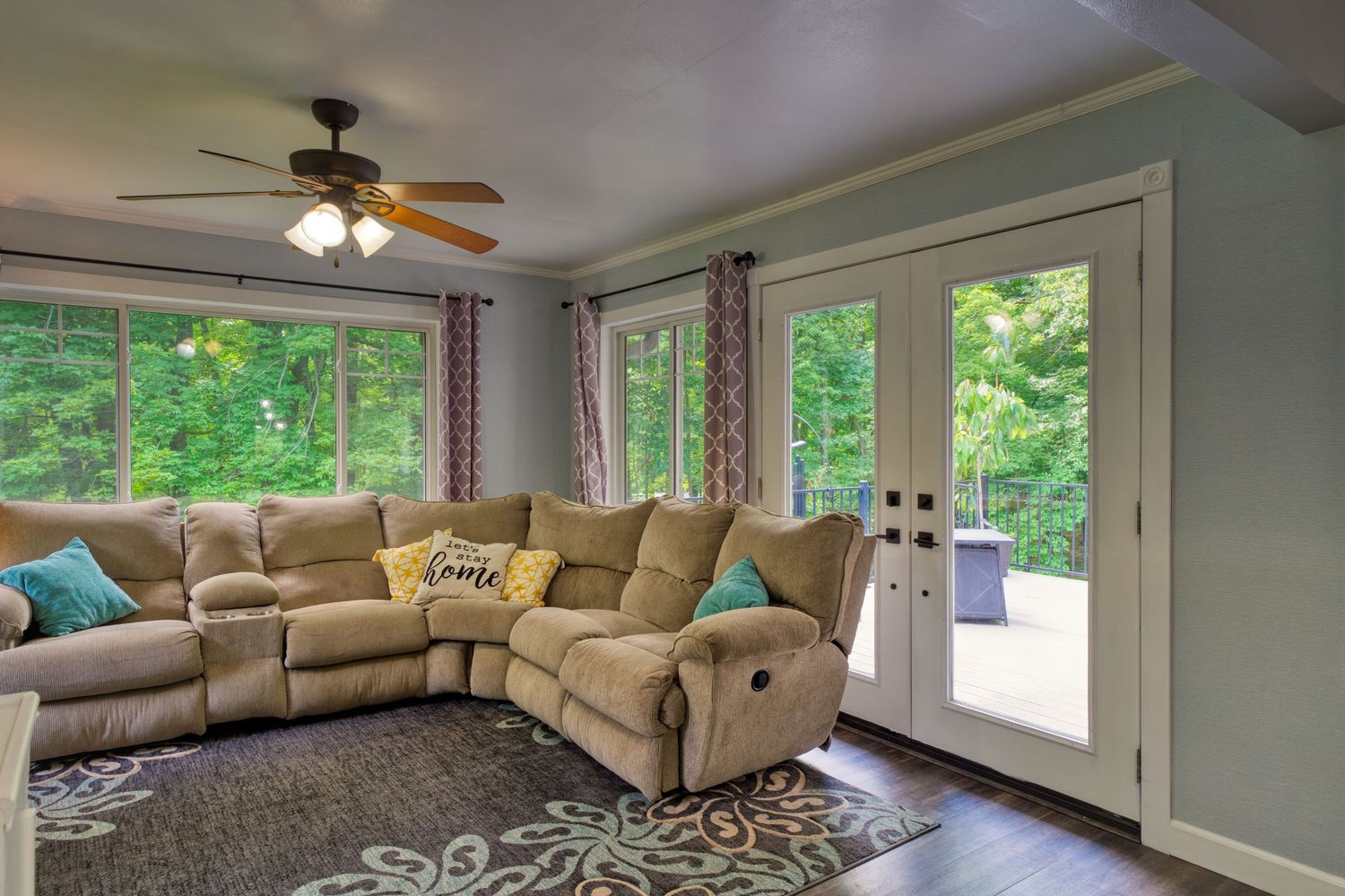 Bright living room with tan sectional sofa, ceiling fan, large windows, French doors, and hardwood floors.