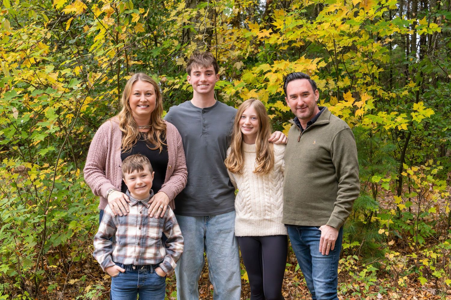 Family of five smiling together outdoors among colorful autumn trees with yellow and green foliage.