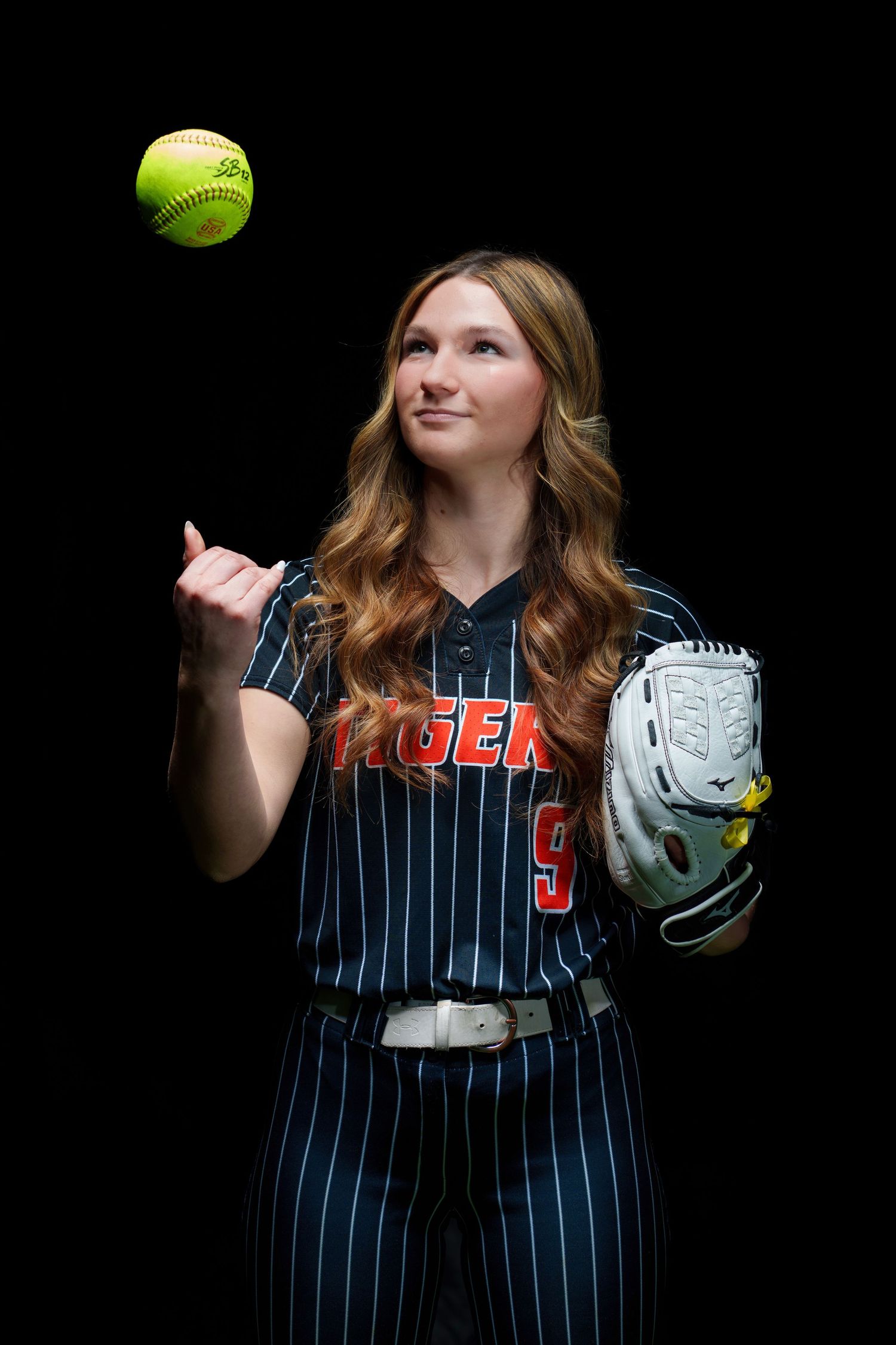 Female softball player in Tigers uniform tossing a yellow softball while holding a Mizuno glove against black background.