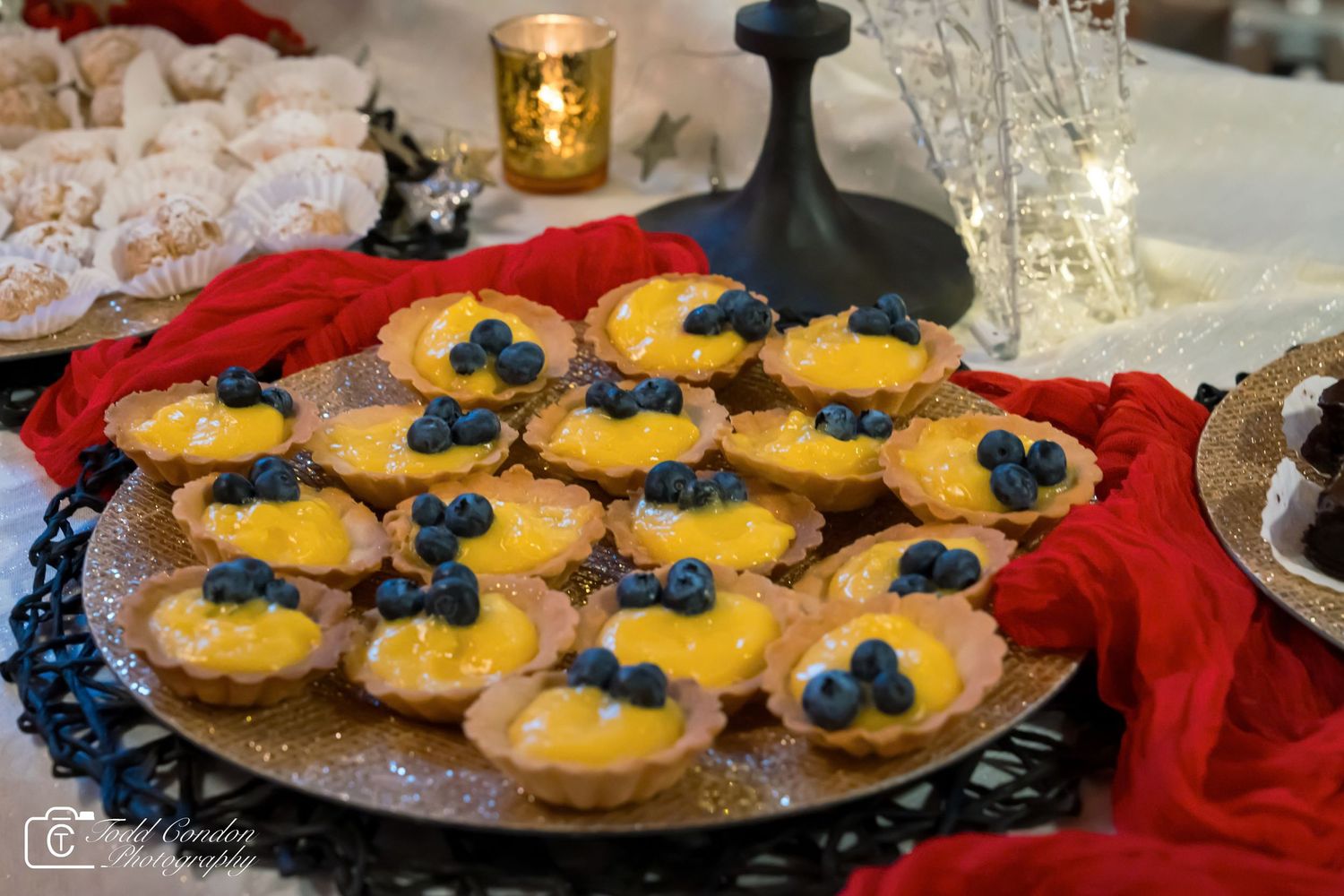Elegant lemon curd tarts topped with fresh blueberries on a decorative plate at a festive holiday dessert table.