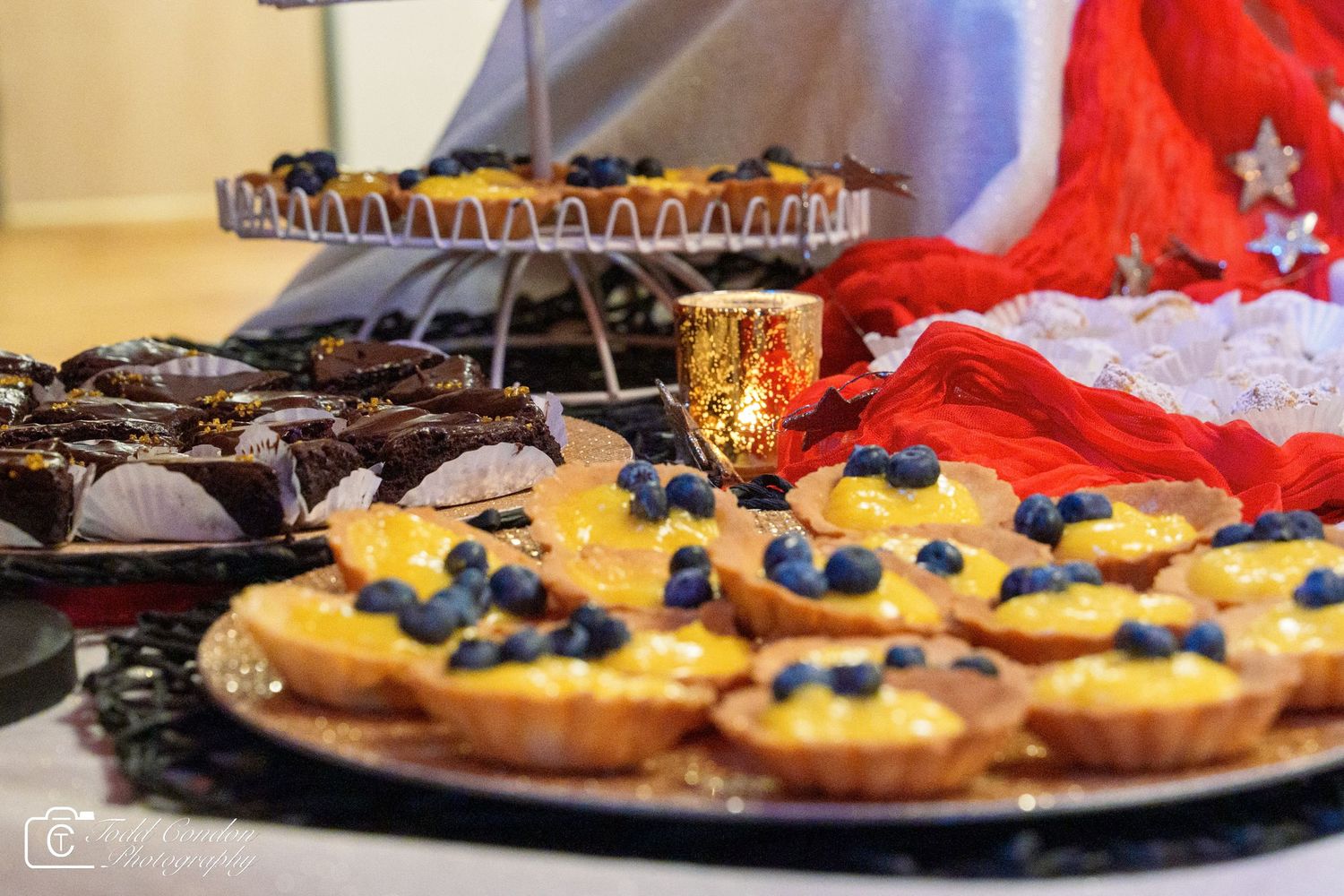 Dessert table with blueberry lemon tarts, chocolate pastries, and mini tarts on a tiered stand at a festive event.