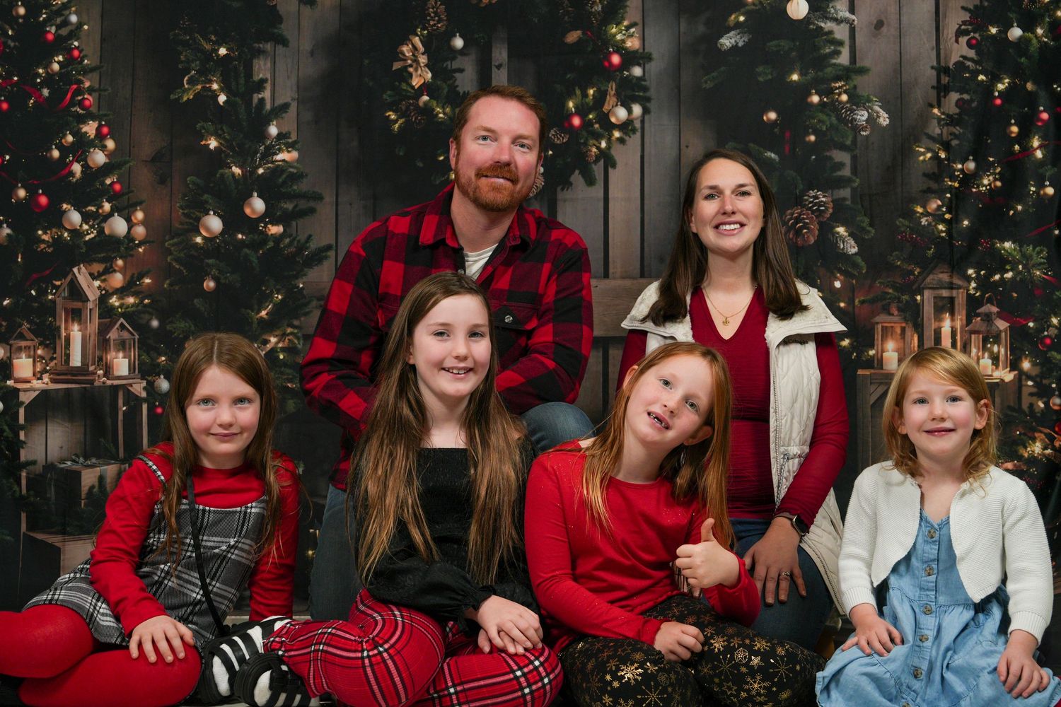 A family poses together in coordinating red and denim outfits in front of decorated Christmas trees.