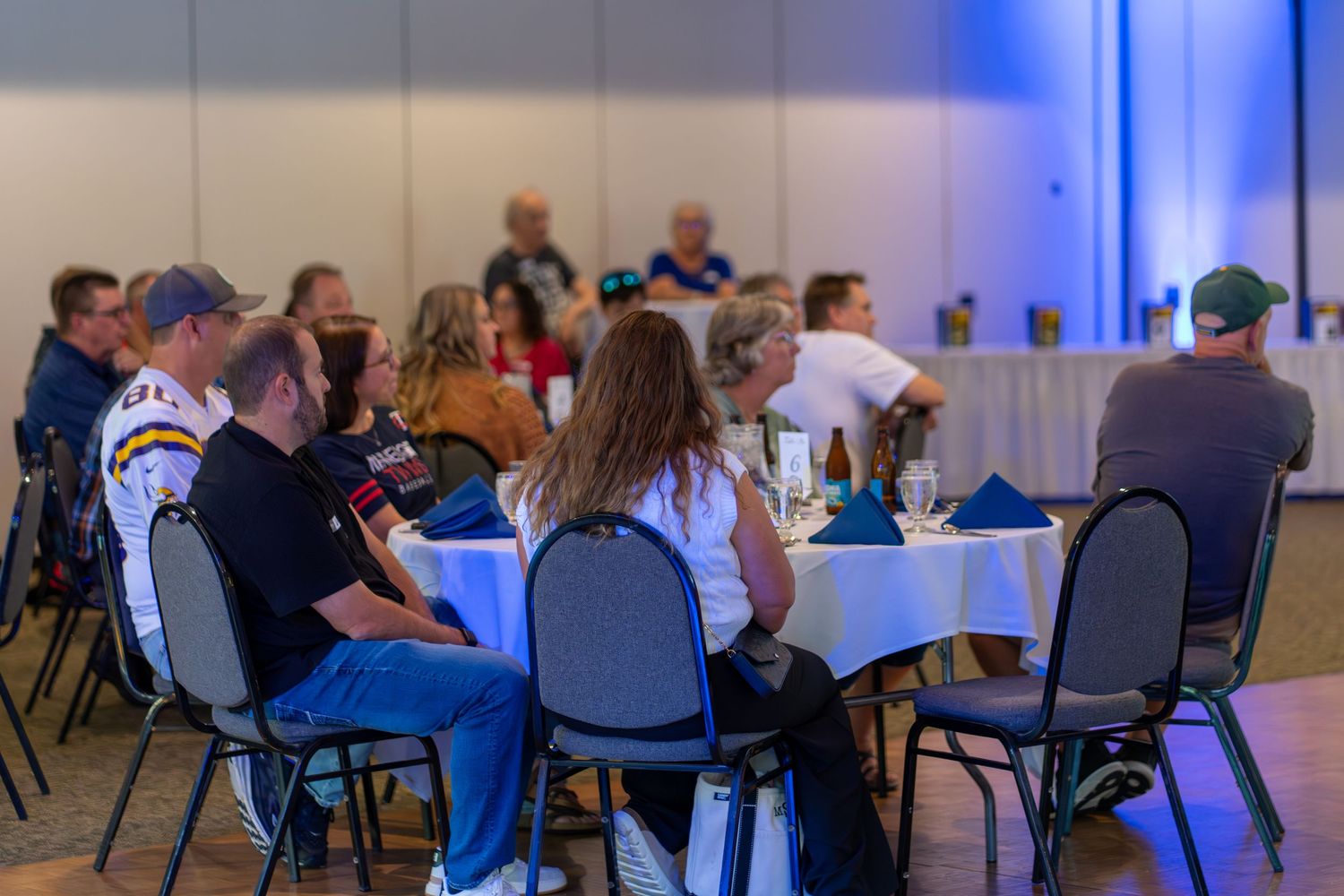 Conference attendees seated at round tables in a large event space with blue ambient lighting.
