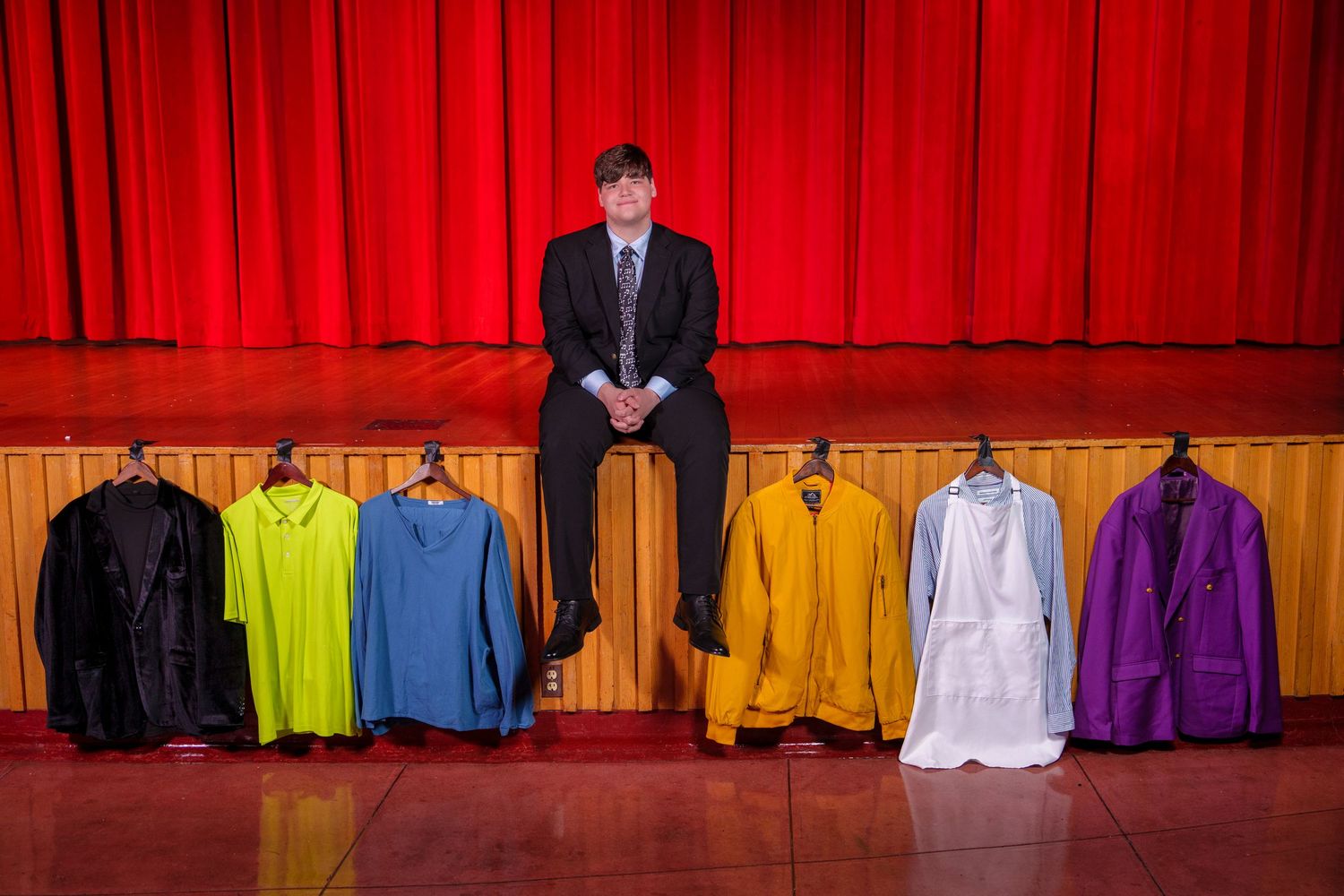 Man in suit sitting on stage behind colorful shirts on hangers with red curtain backdrop.