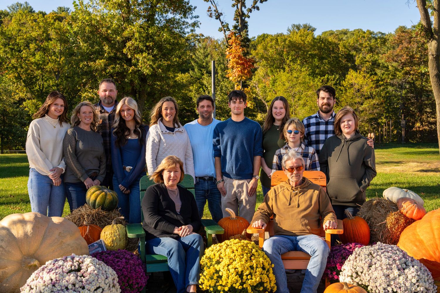 Family group photo outdoors in autumn with pumpkins and mums, surrounded by fall foliage and colorful decorations.