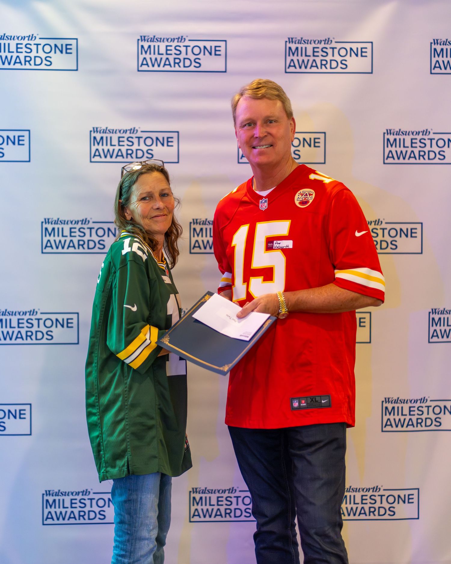 Two sports fans in opposing team jerseys pose together at an awards ceremony backdrop with Milestone Awards logos.