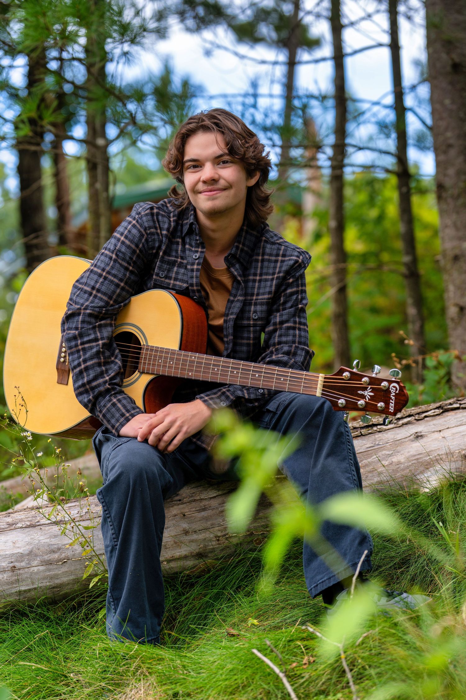 A musician sits on a fallen log in a forest setting while holding an acoustic guitar among pine trees and greenery.