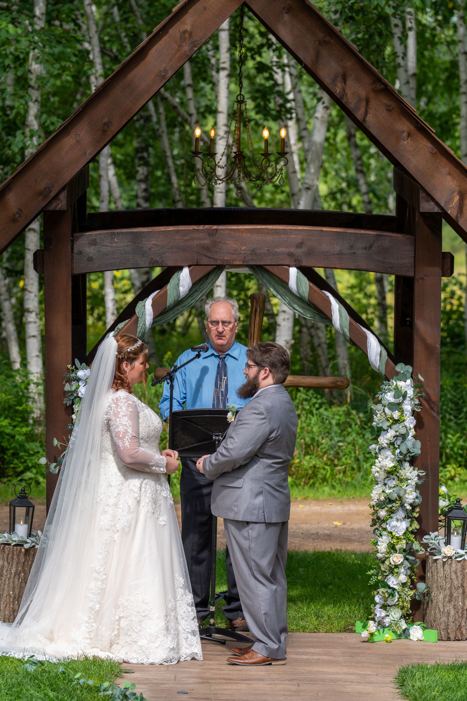 Wedding ceremony takes place under a rustic wooden pavilion surrounded by birch trees in a forest setting.