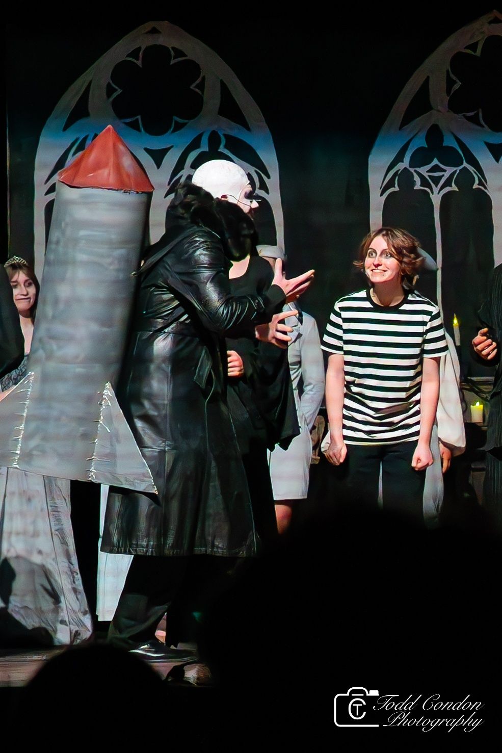 Theatrical performance with actors in gothic arches setting showing someone in black coat and a person in striped shirt.