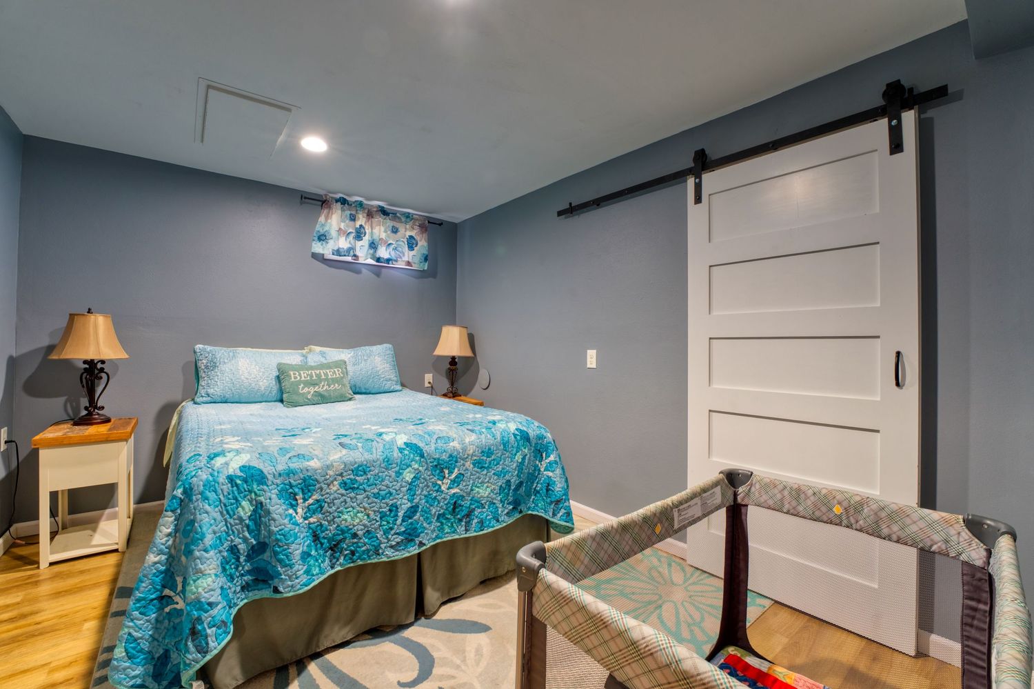 Cozy bedroom with teal floral bedspread, barn door, hardwood floors, and soft lamp lighting in a blue-walled room.