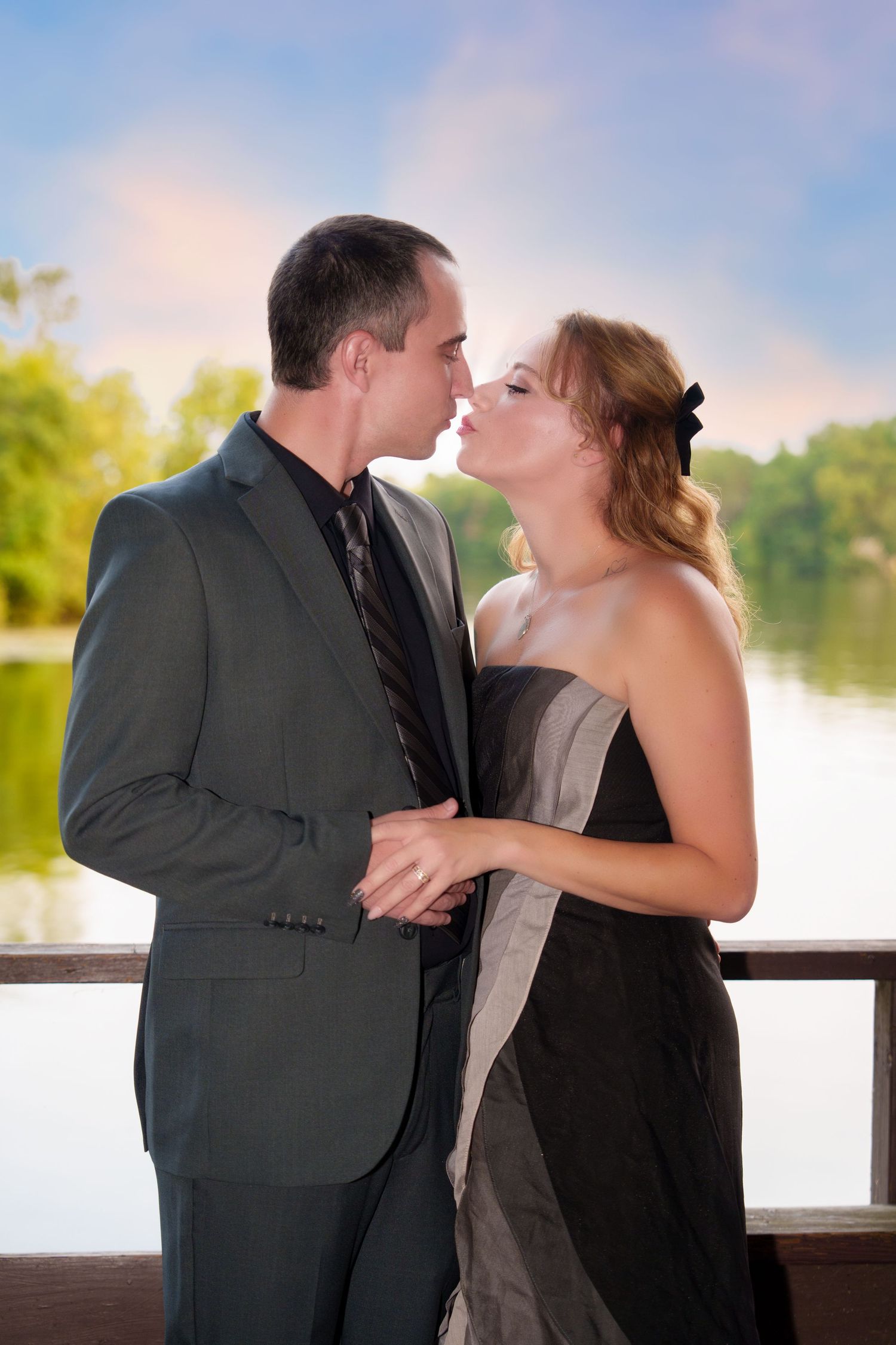 Couple in formal attire sharing a romantic moment on a lakeside deck, man in gray suit and woman in black dress.