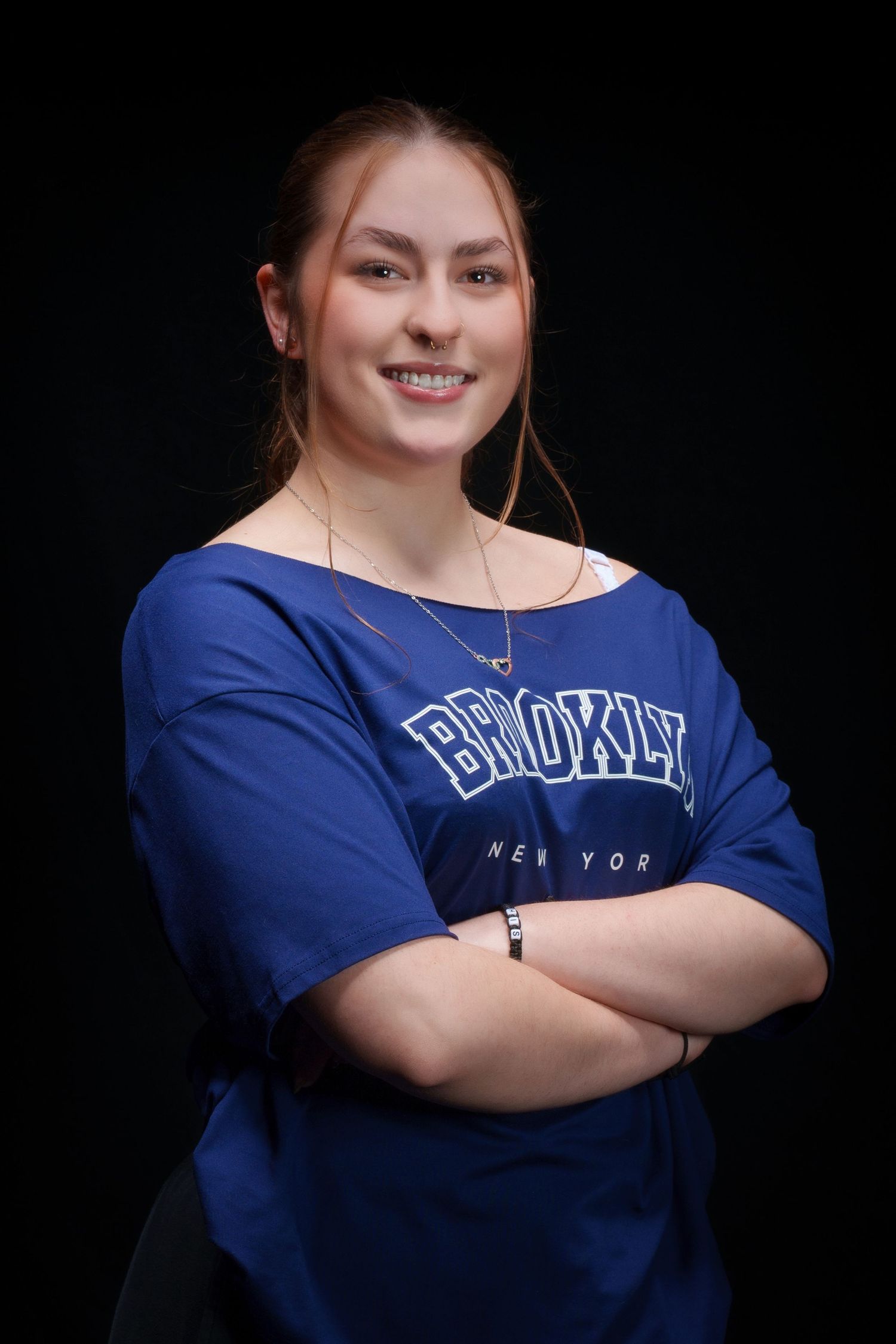 Smiling young woman in blue Brooklyn New York shirt standing with arms crossed against black background.