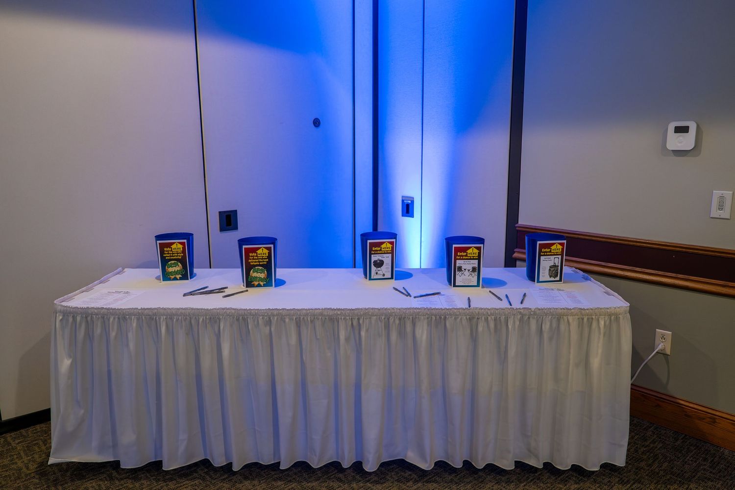 Blue uplighting illuminates a white-draped banquet table setup against a wall in an event space.