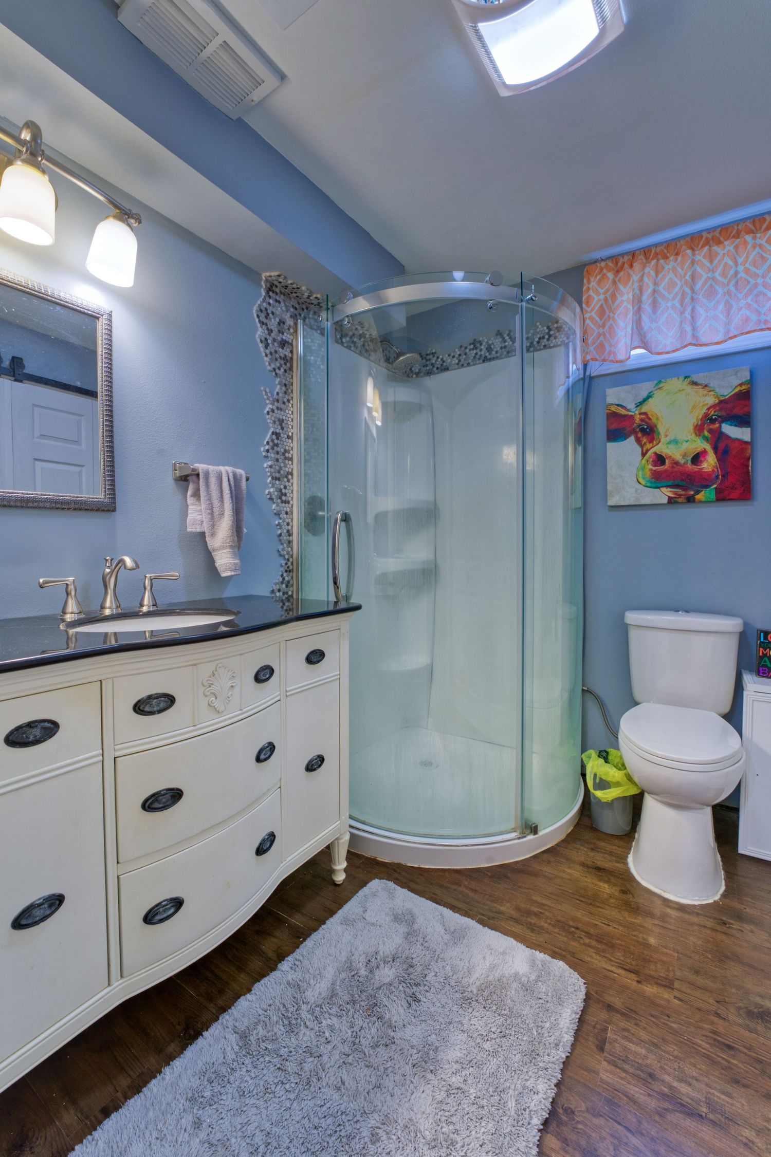 Blue bathroom with curved glass shower, vintage white dresser vanity, hardwood floors, and colorful cow painting above toilet.