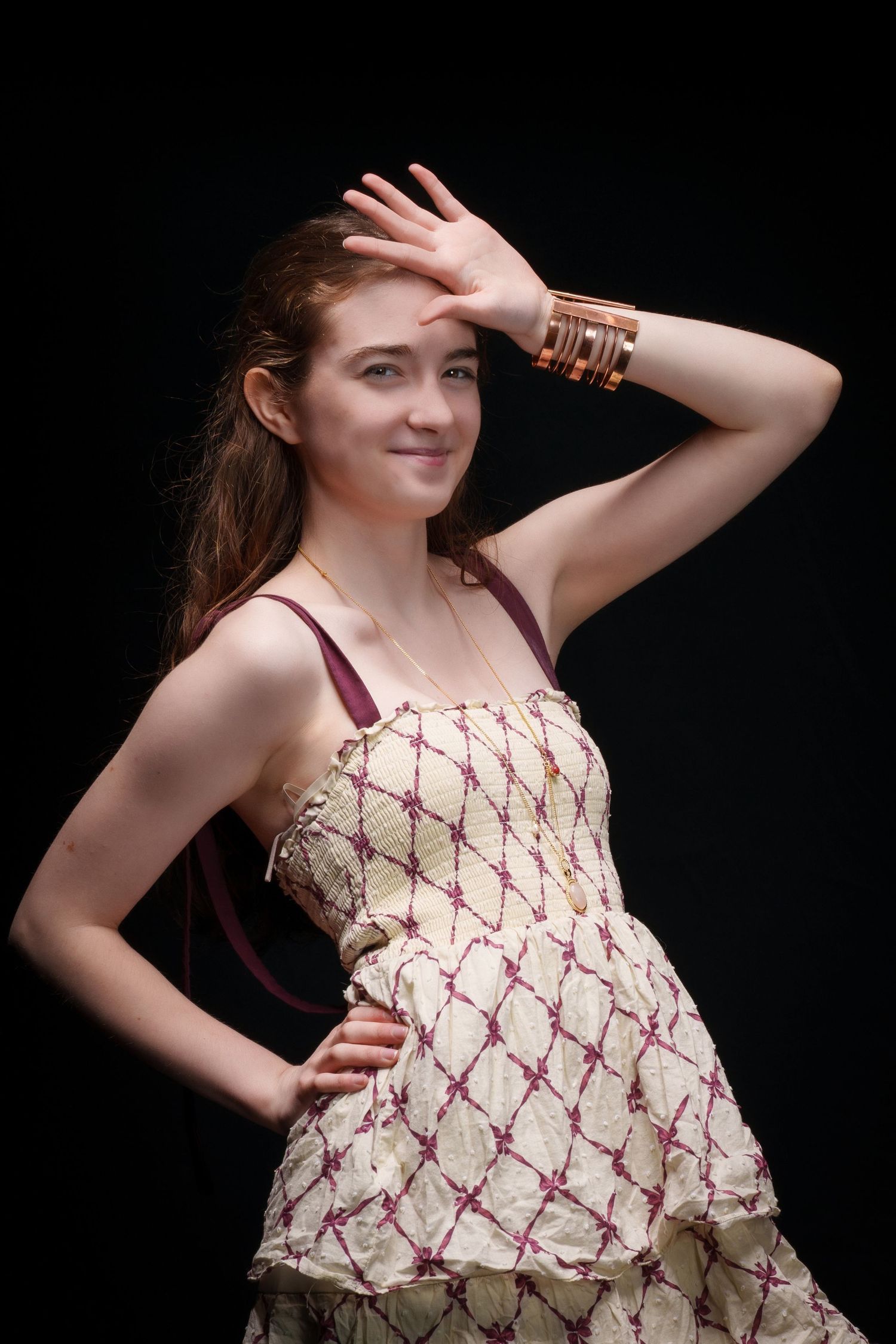 Young woman in cream and purple dress poses with hand raised, wearing gold bangles, against black background.