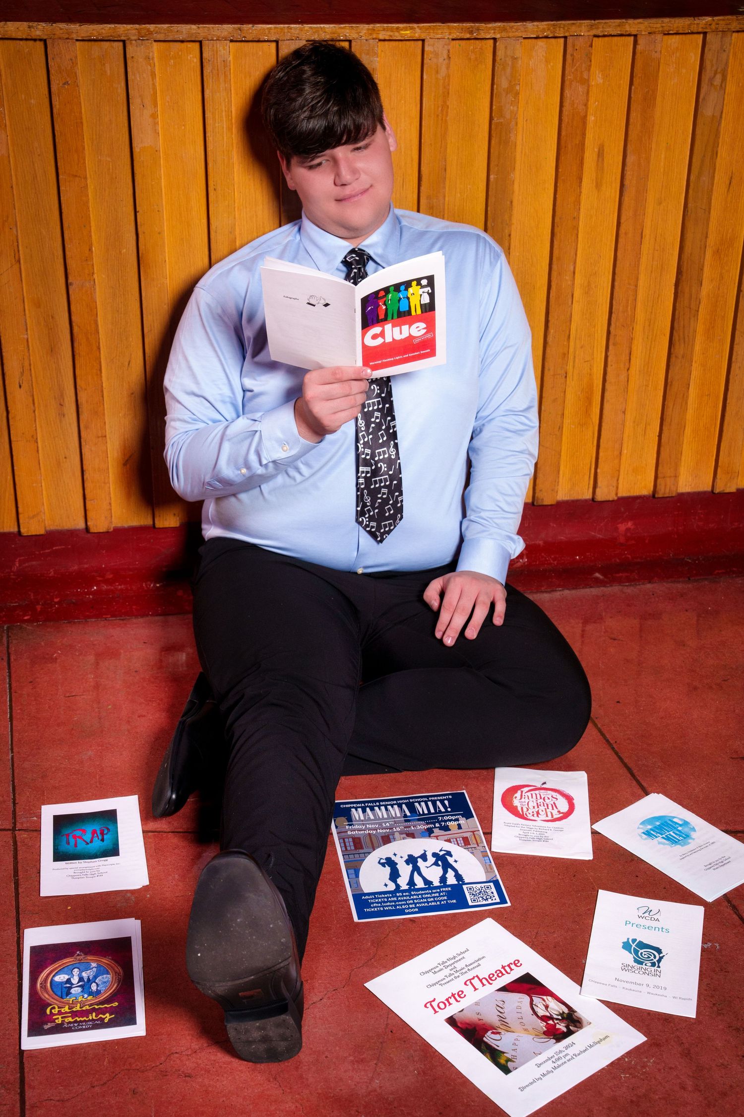 Young man in dress shirt and tie sits on floor reading Clue book, surrounded by theater program pamphlets.