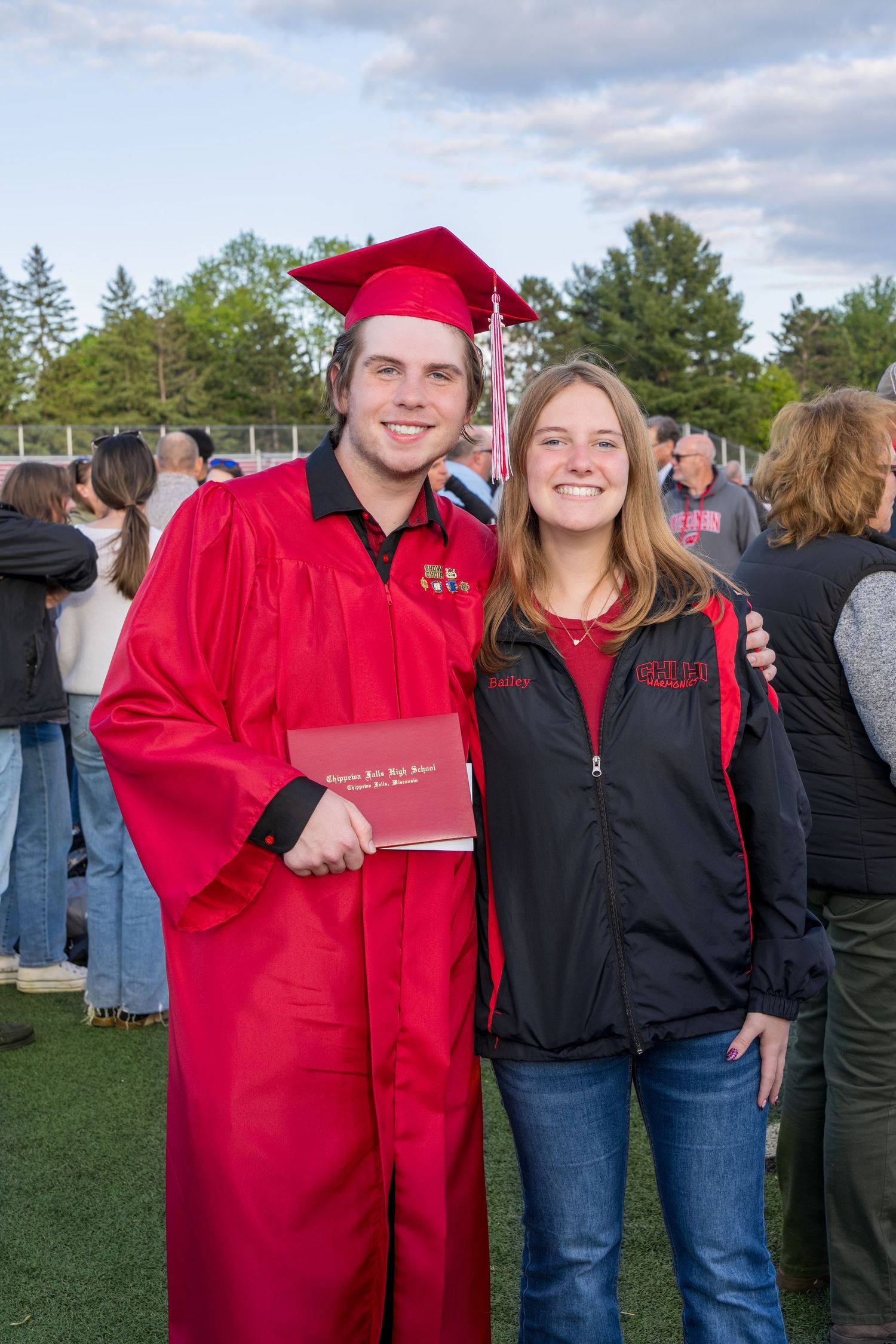 A smiling graduate in red cap and gown holds diploma beside a young woman outdoors at graduation ceremony.