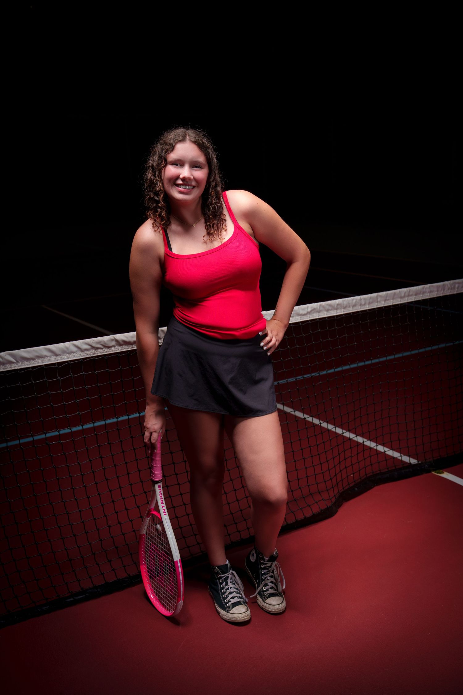 Smiling teenage girl in red top and black skirt holding pink tennis racket while posing by net on indoor court.