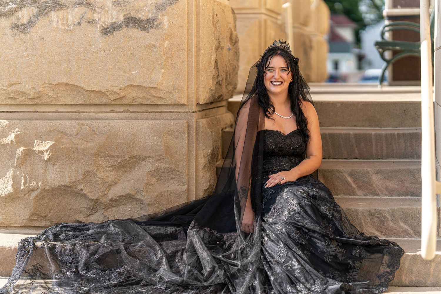 A person in a flowing black gown poses elegantly on stone steps in an outdoor setting.