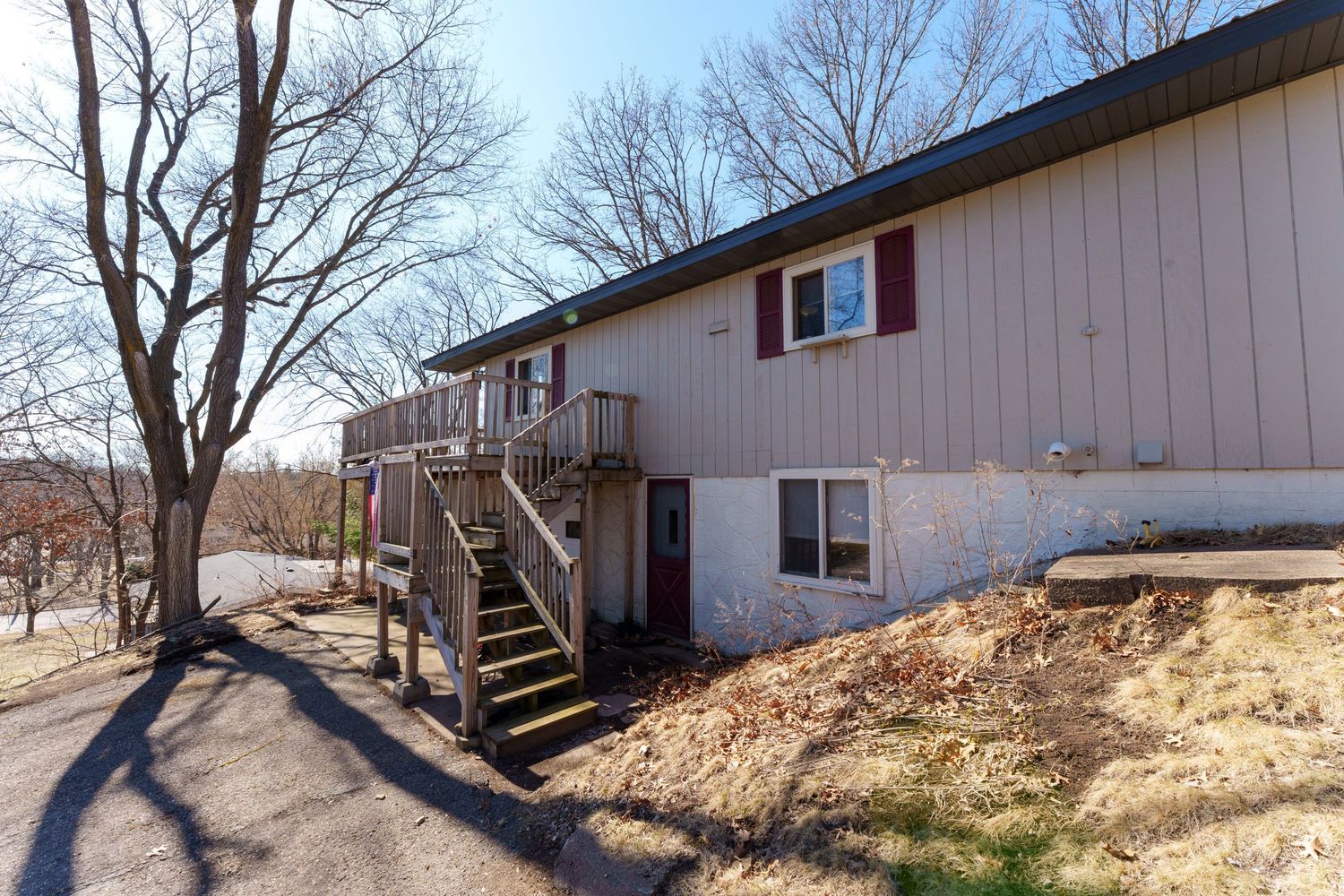 Long white mobile home with wooden deck and stairs sits on a sloping lot surrounded by bare trees in winter.