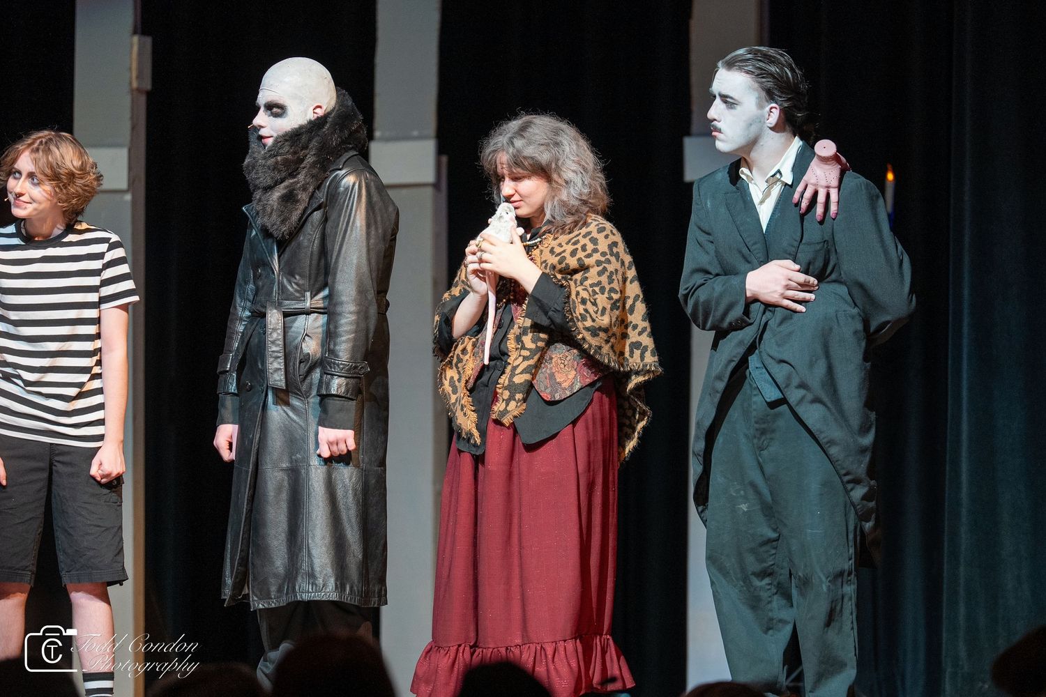 A group of performers in period costumes stand on a dark stage during a theatrical production.