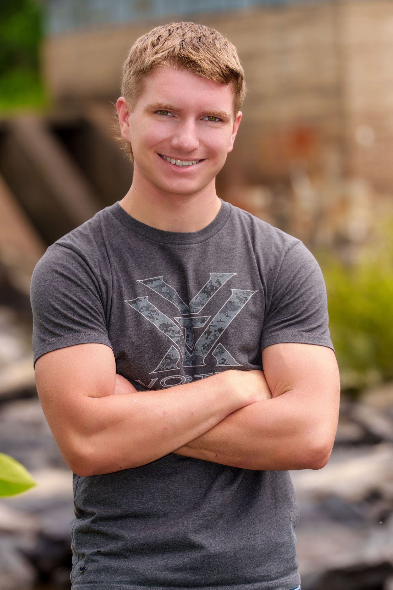 Smiling young man with blonde hair, arms crossed, wearing a gray graphic t-shirt, posing outdoors.
