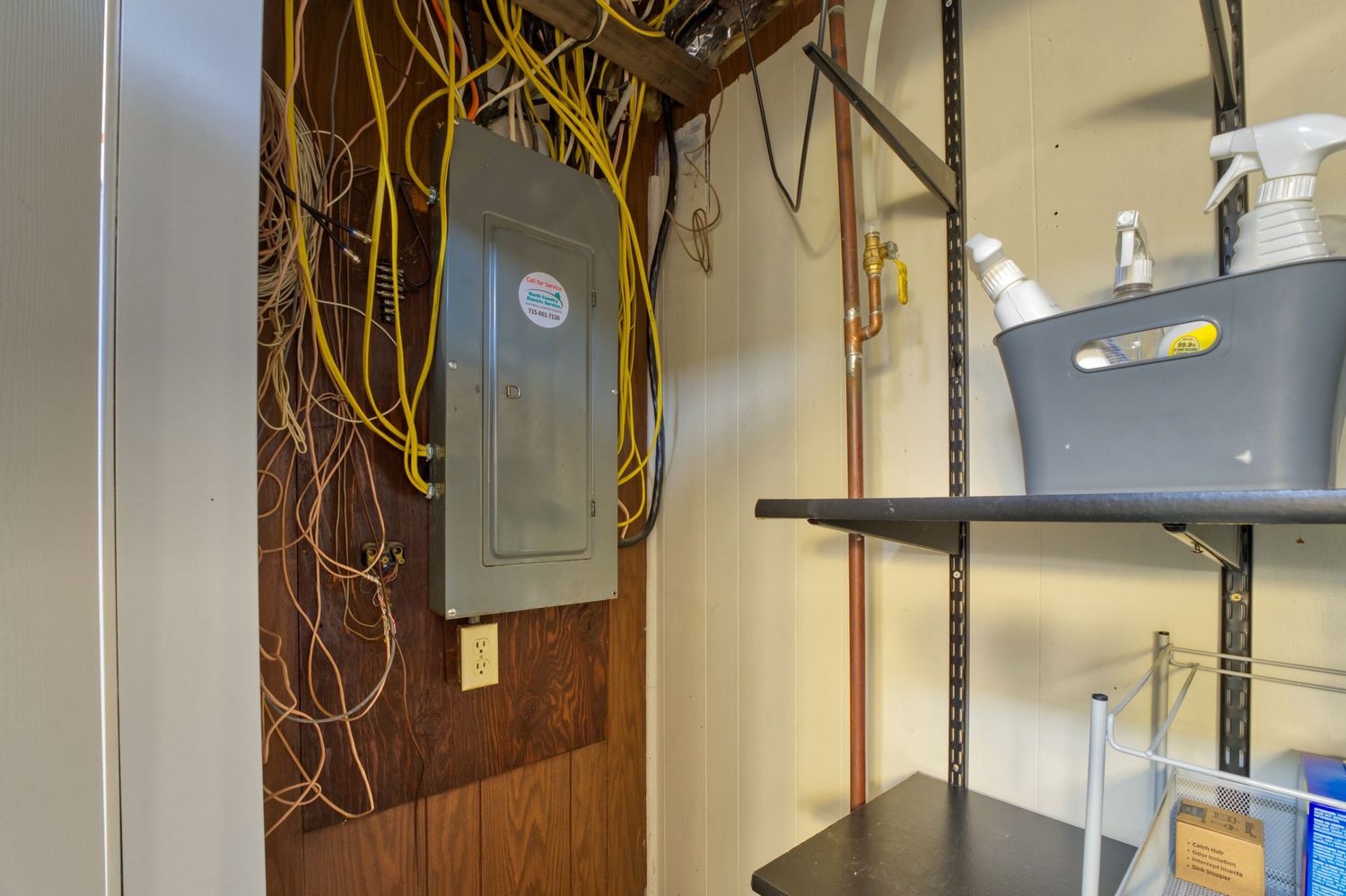 Utility room with electrical panel, yellow wiring, metal shelving, and cleaning supplies on white walls.