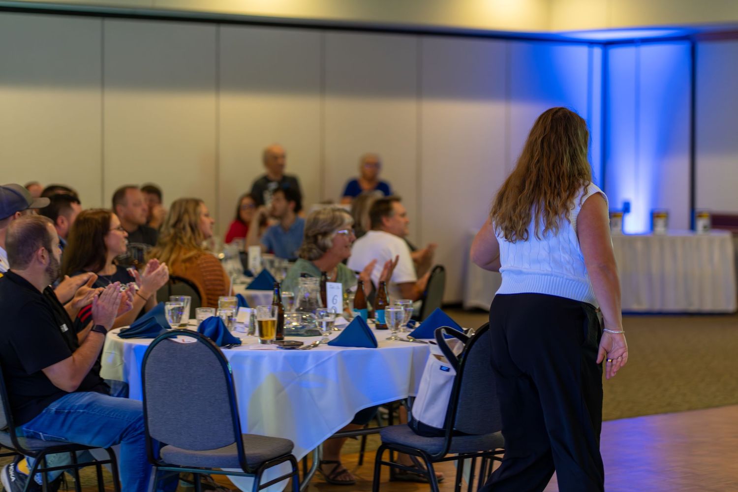 People seated at round tables in a large conference or event room with blue lighting accents.