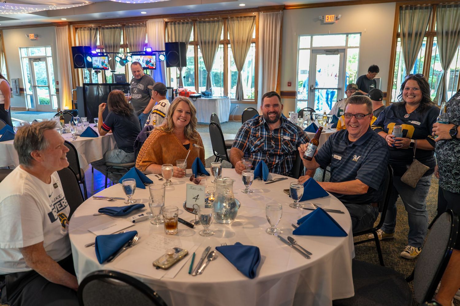 People gather at round tables in a banquet hall for a social event with blue and white decor.