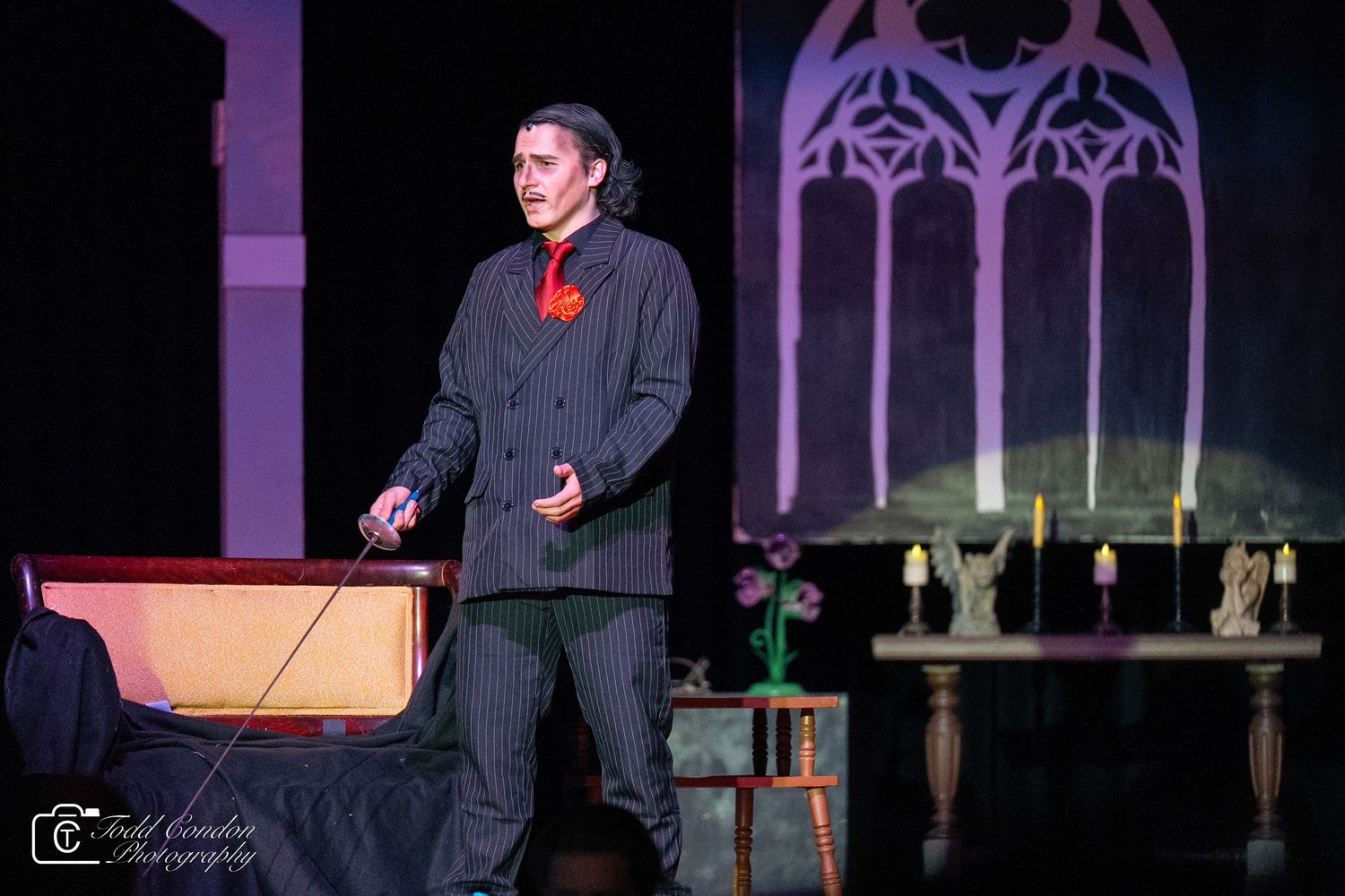 A performer stands on stage during a theatrical production with dramatic purple lighting and set pieces.