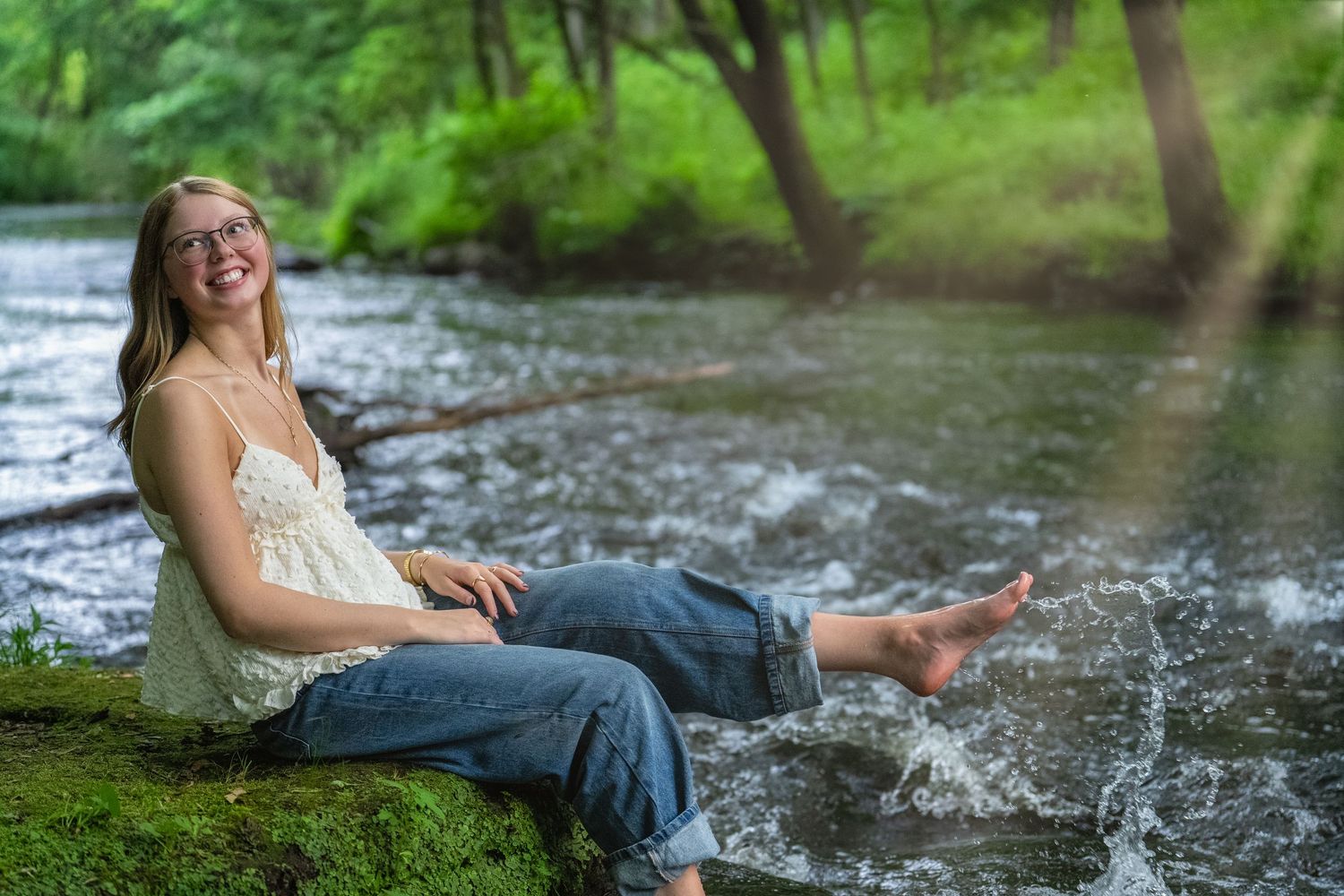 Someone sits on mossy riverbank wearing jeans and white top while splashing feet in flowing stream on sunny day.
