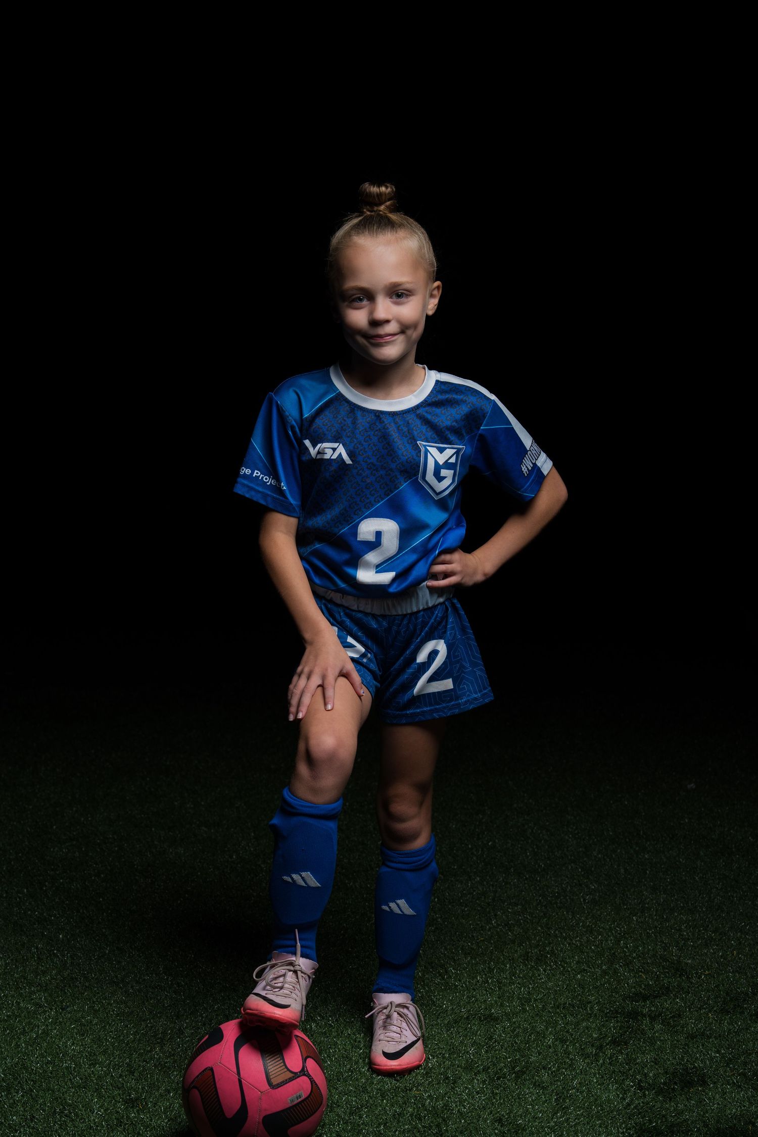 Young athlete in blue soccer uniform stands with pink soccer ball on indoor turf field.