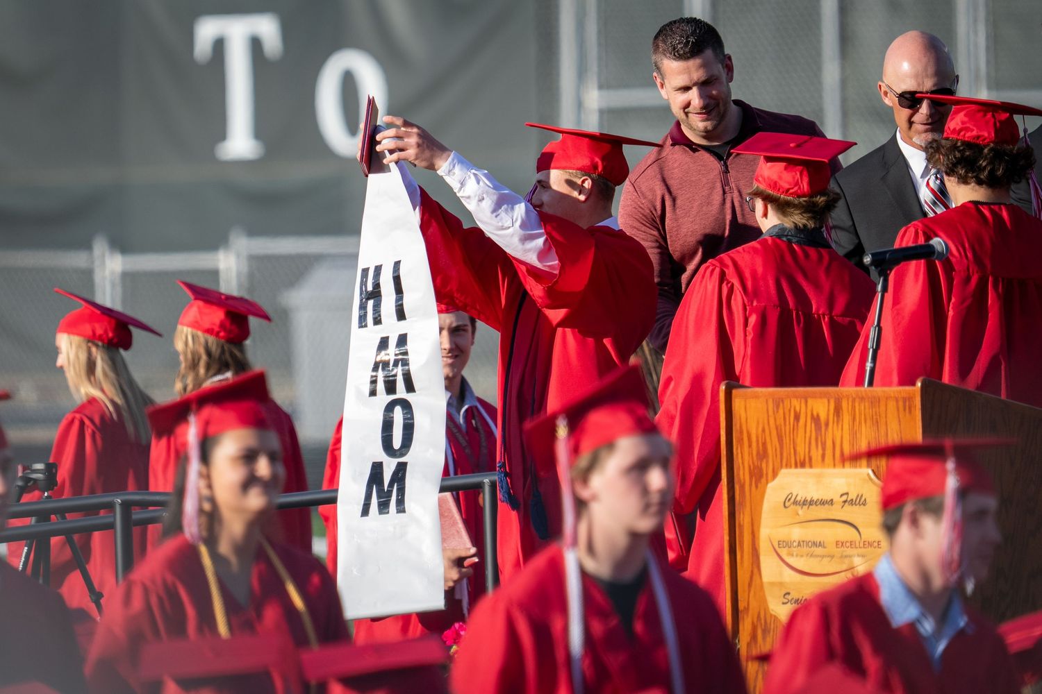 Graduates in red caps and gowns at commencement ceremony, one holding a white 'Hi Mom' banner on stage.