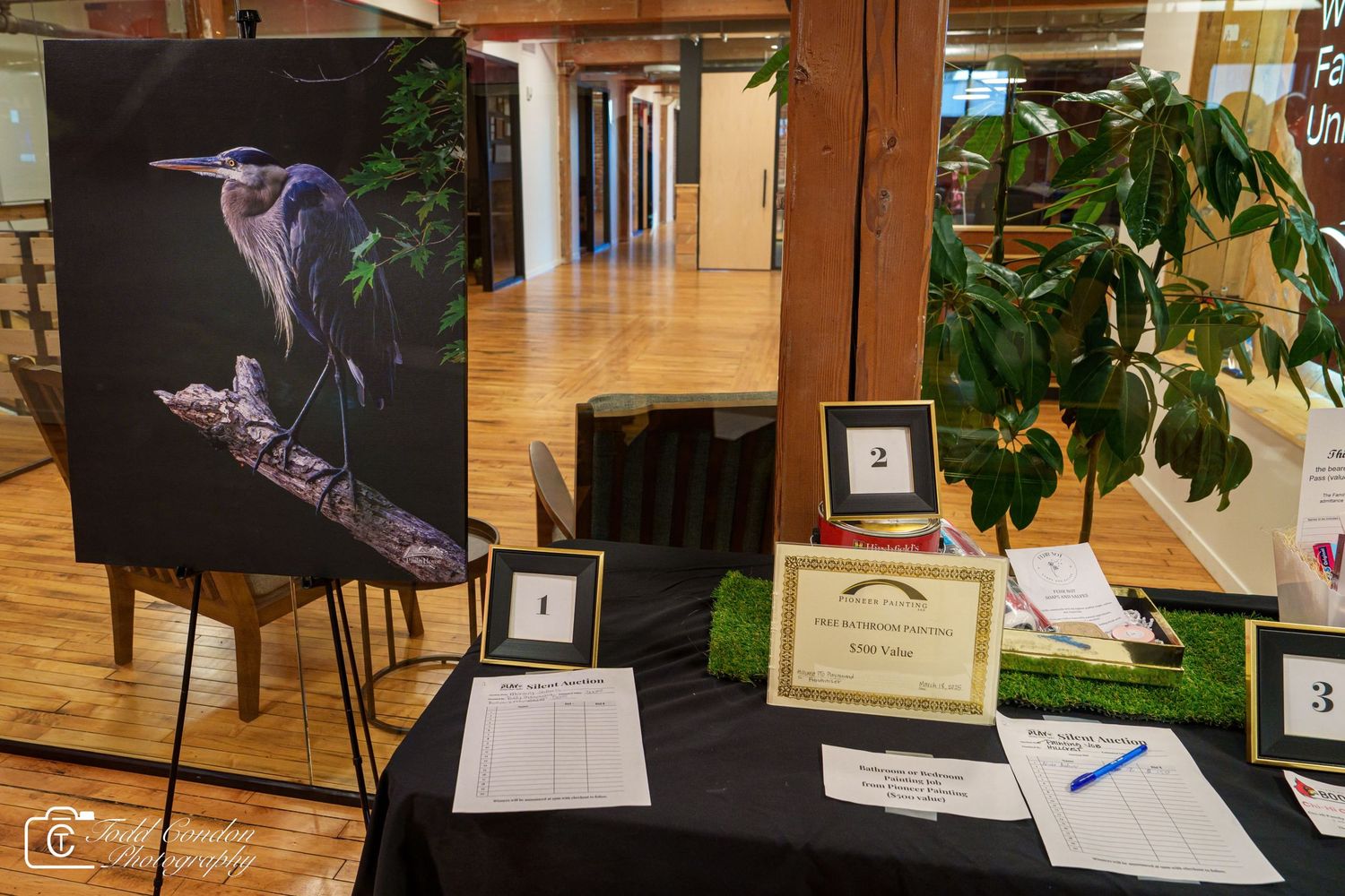 Art auction display featuring a heron painting on easel with bidding sheets and numbered lots on black draped table.