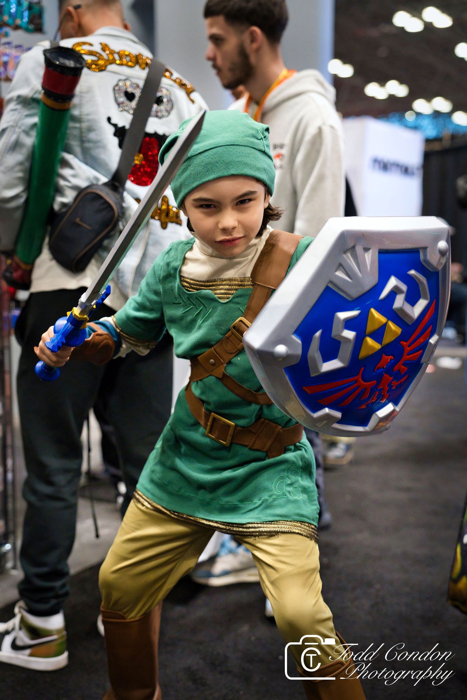 Child cosplaying as Link from Legend of Zelda, holding sword and Hylian Shield at a gaming convention.