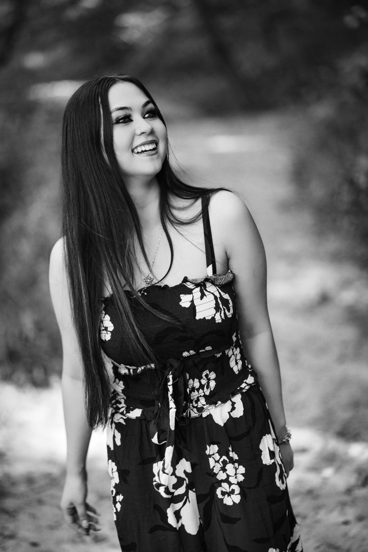 Black and white portrait of a smiling young woman with long dark hair wearing a floral dress outdoors.