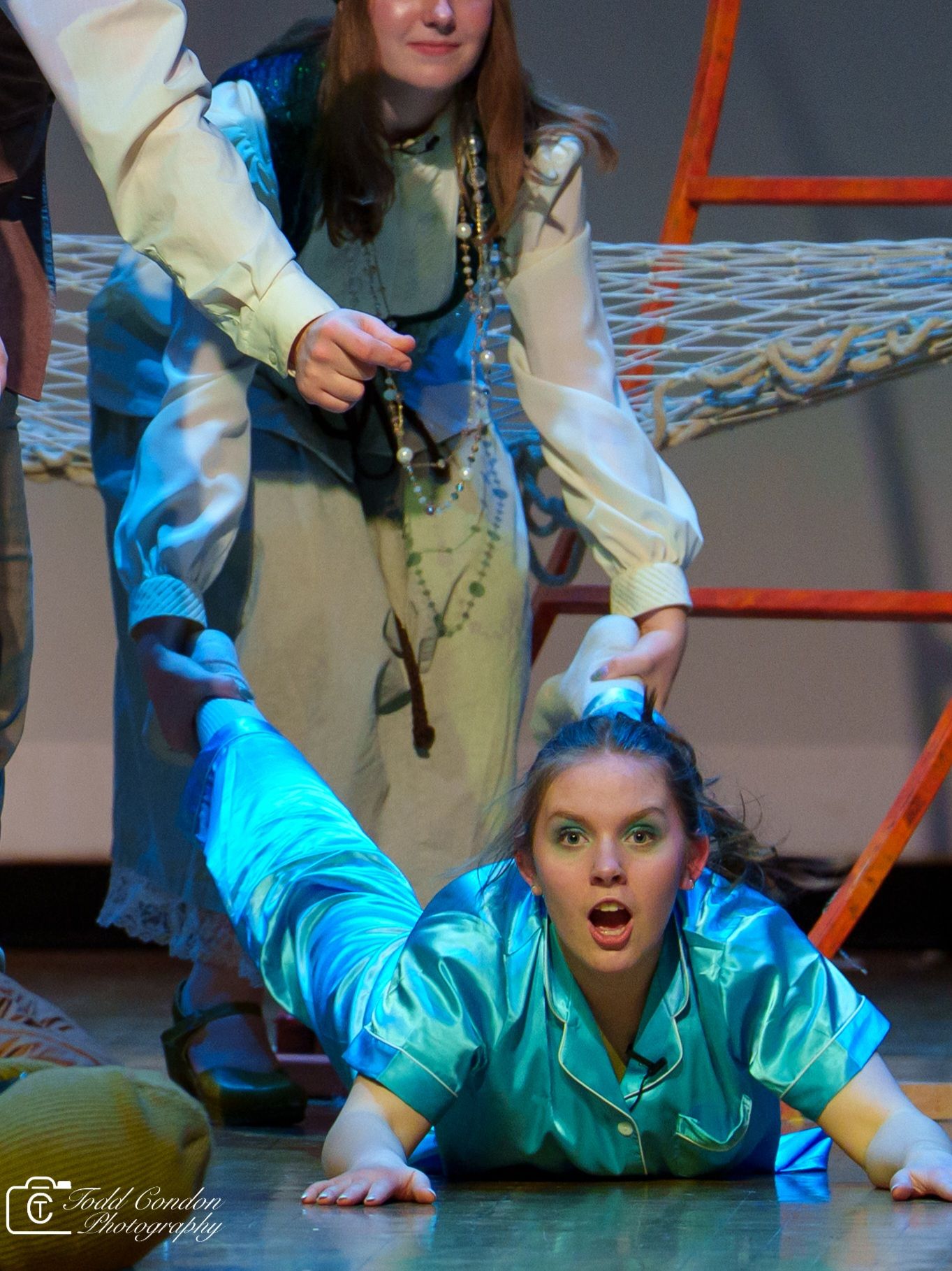 Performers in theatrical costumes act out a dramatic scene on stage with one in a turquoise outfit lying down.
