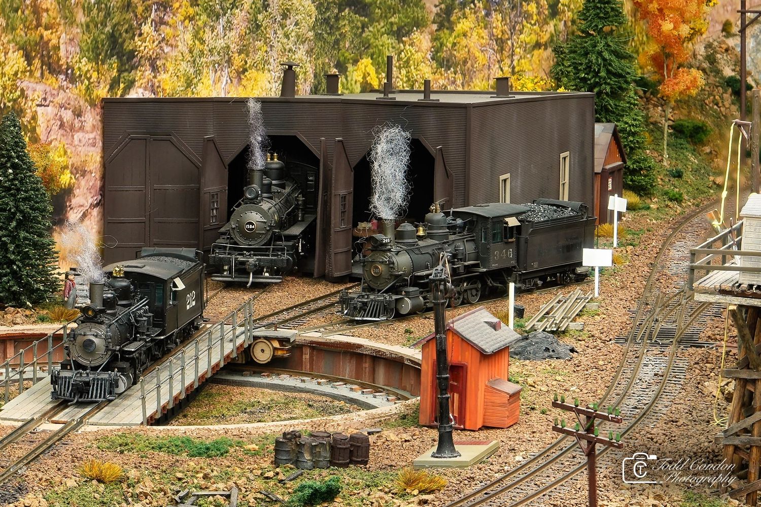 A model train runs through a scenic autumn landscape with colorful fall foliage and miniature buildings along the track.