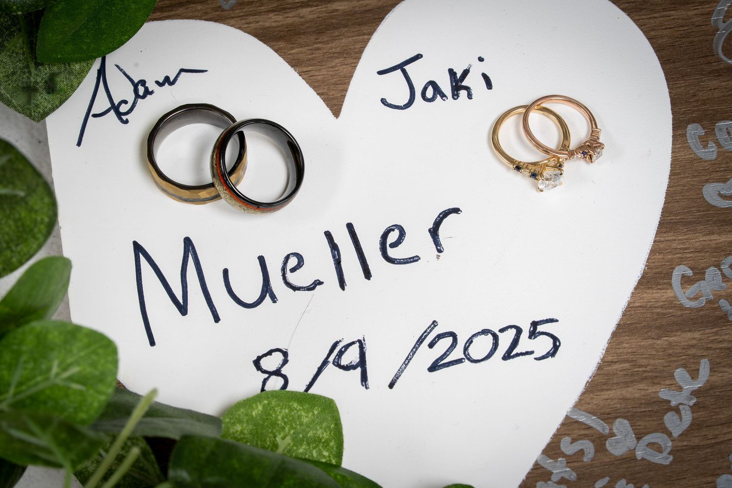 White heart-shaped paper notes with handwriting and wedding rings displayed on a wooden surface with green foliage.
