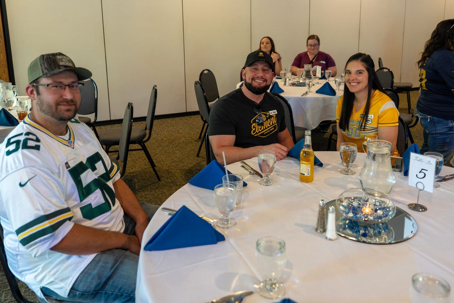 Guests seated at a round table with drinks during a casual dining event, with one wearing a Green Bay Packers jersey.