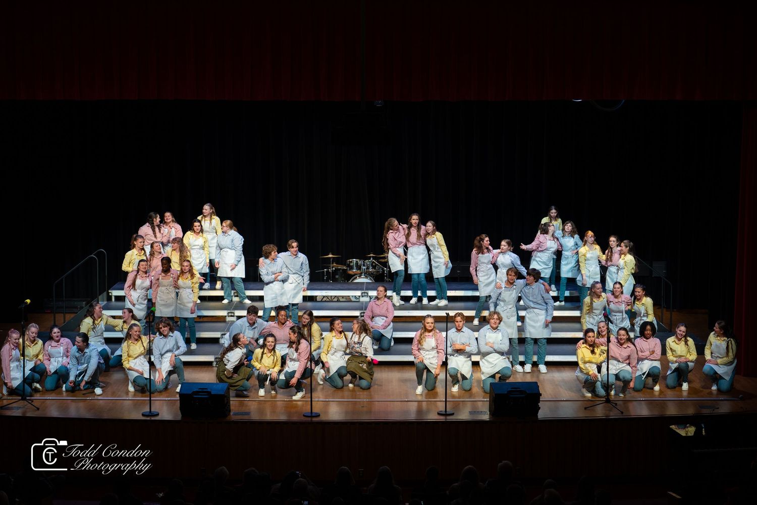 Stage performance featuring multiple performers seated in a row wearing colorful casual attire against a dark backdrop.