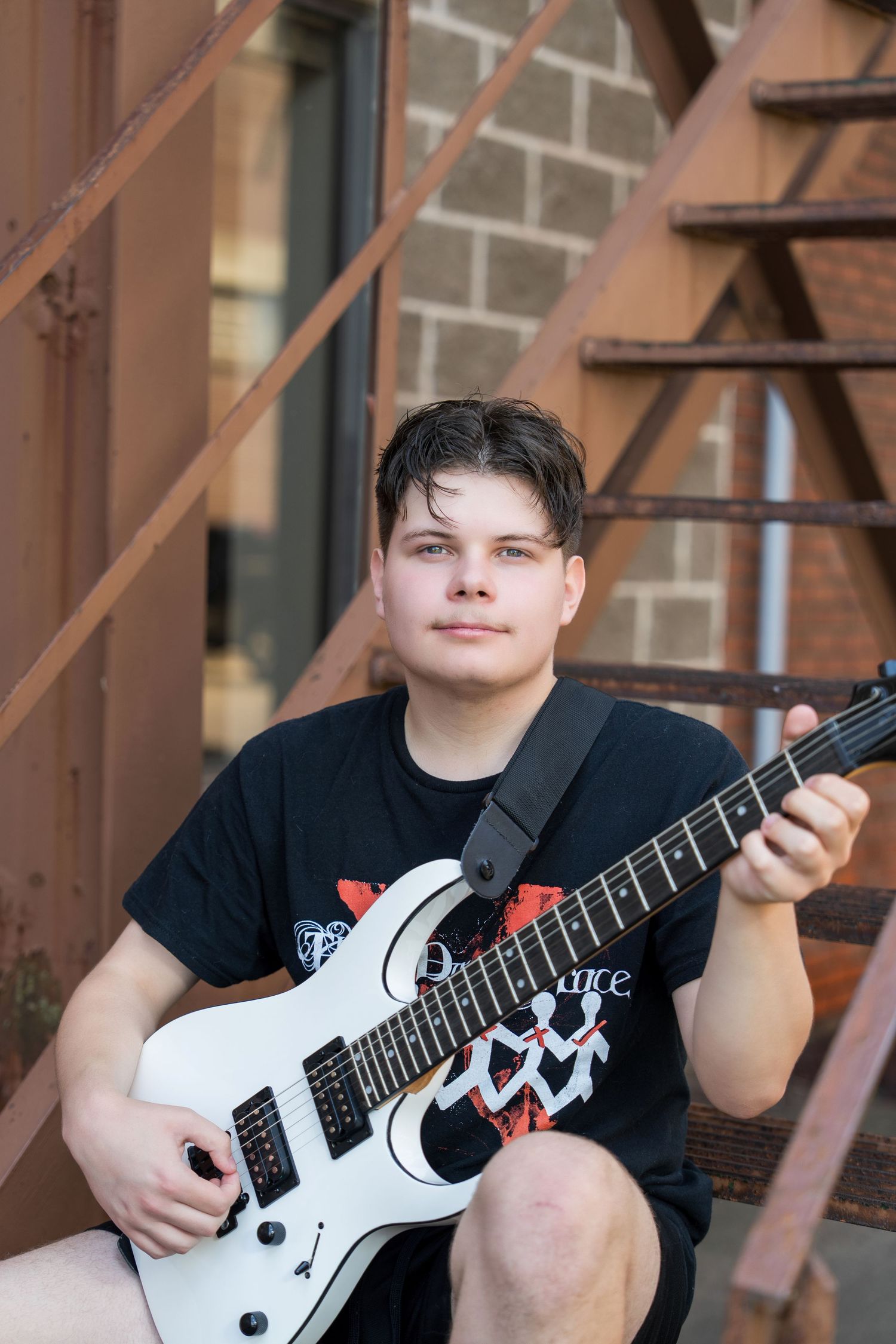 A musician in a black t-shirt plays a white electric guitar against a brick wall background.