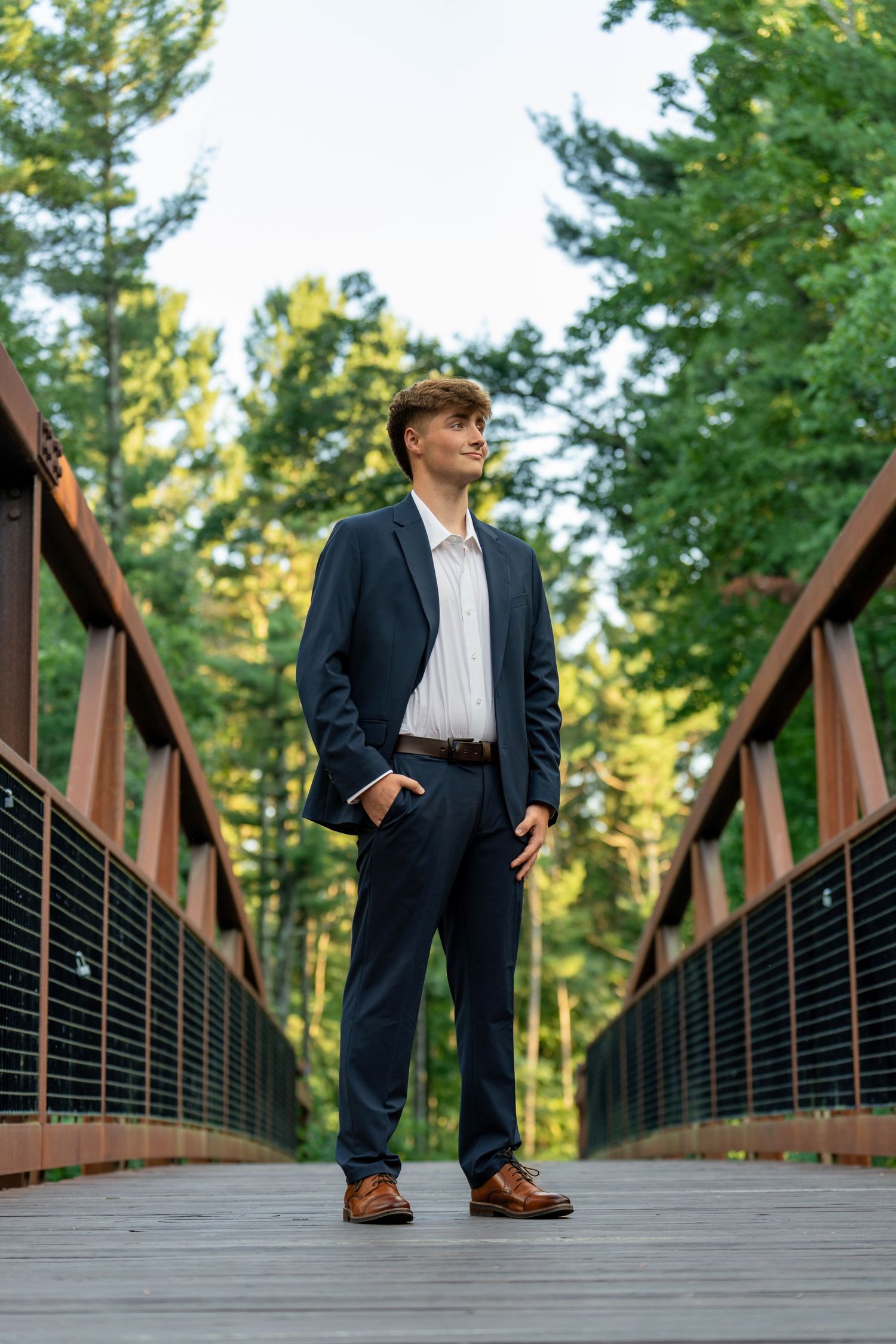 A person in a business suit stands on a wooden bridge surrounded by lush green forest scenery.