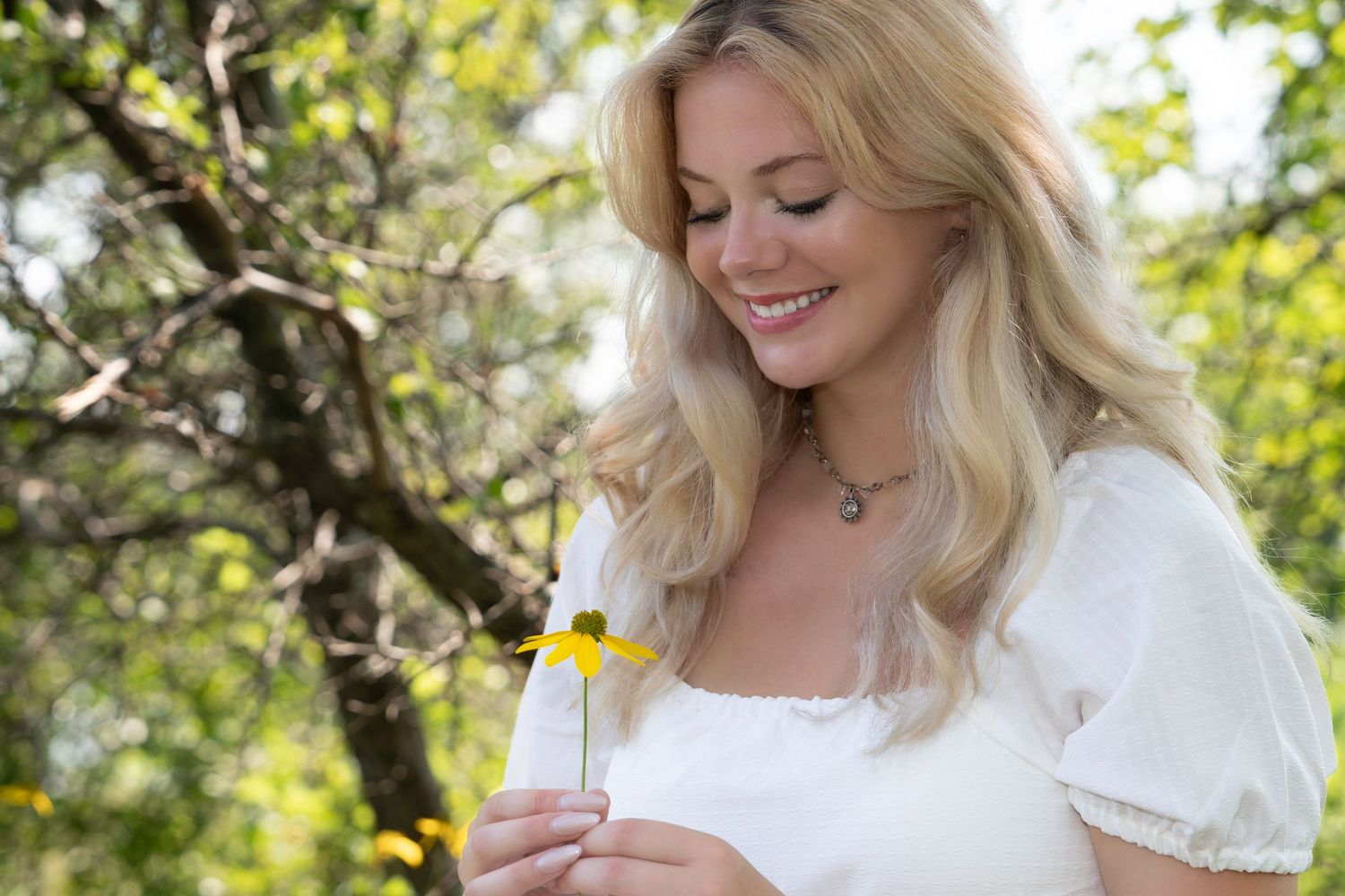 Woman in white dress smiles while holding a bottle of skincare product in a sunny garden setting.