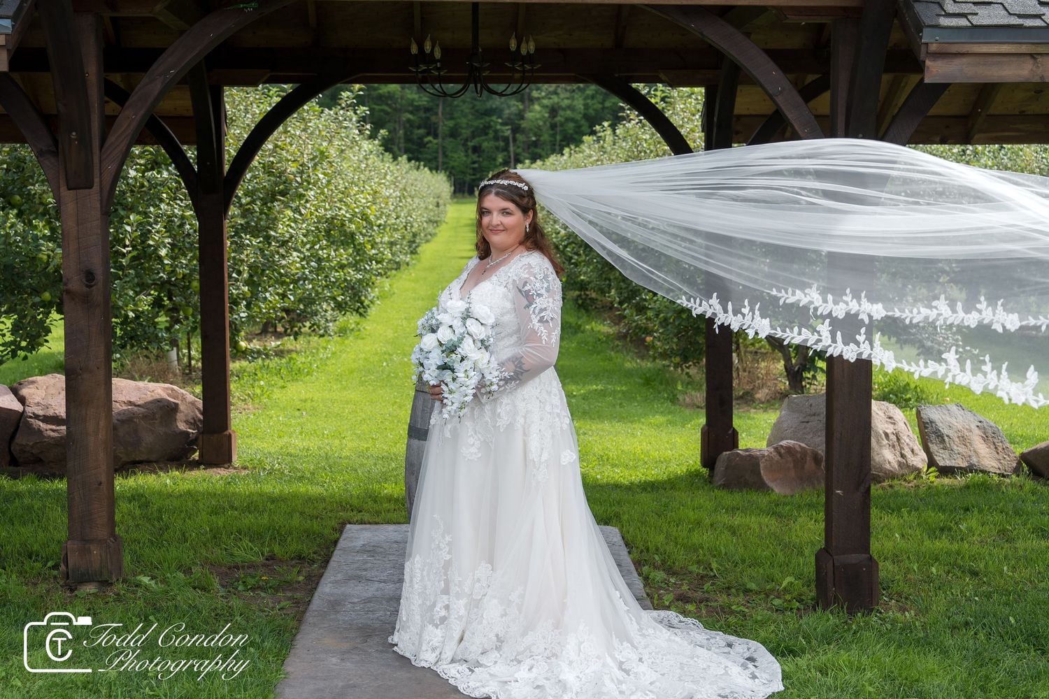 A flowing white wedding veil billows in the breeze beneath a rustic wooden gazebo in a lush garden setting.