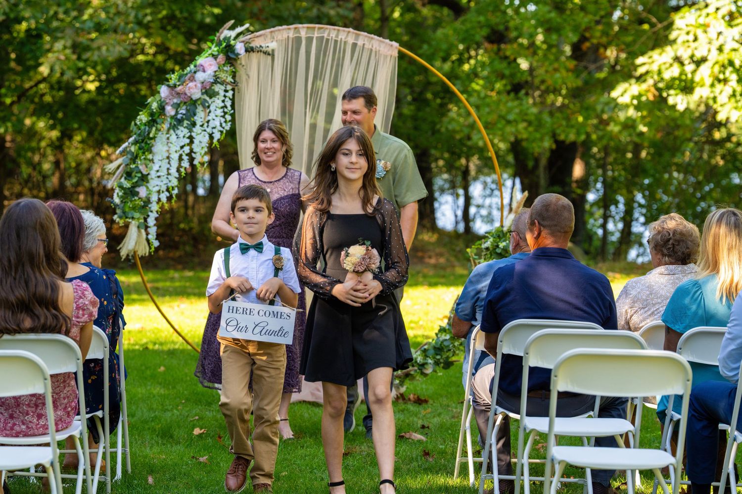Outdoor wedding reception in a garden setting with white chairs and tables arranged under trees.